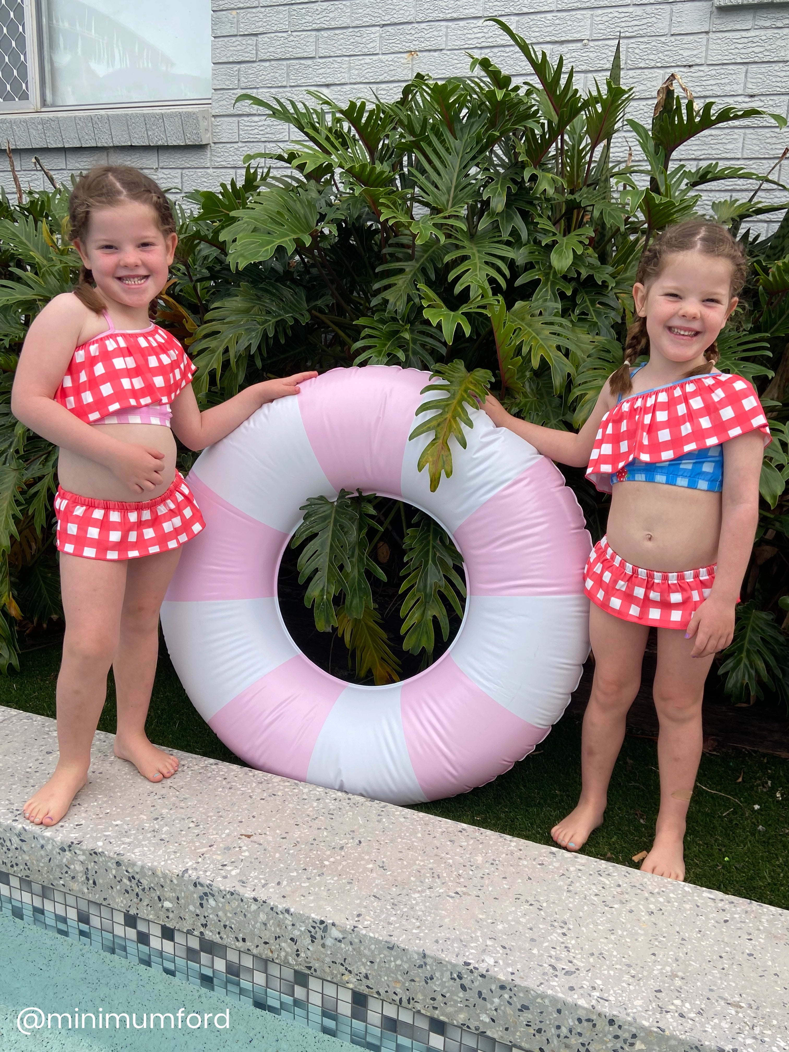 Two children in matching swimsuits with a pink and white inflatable ring by a pool.