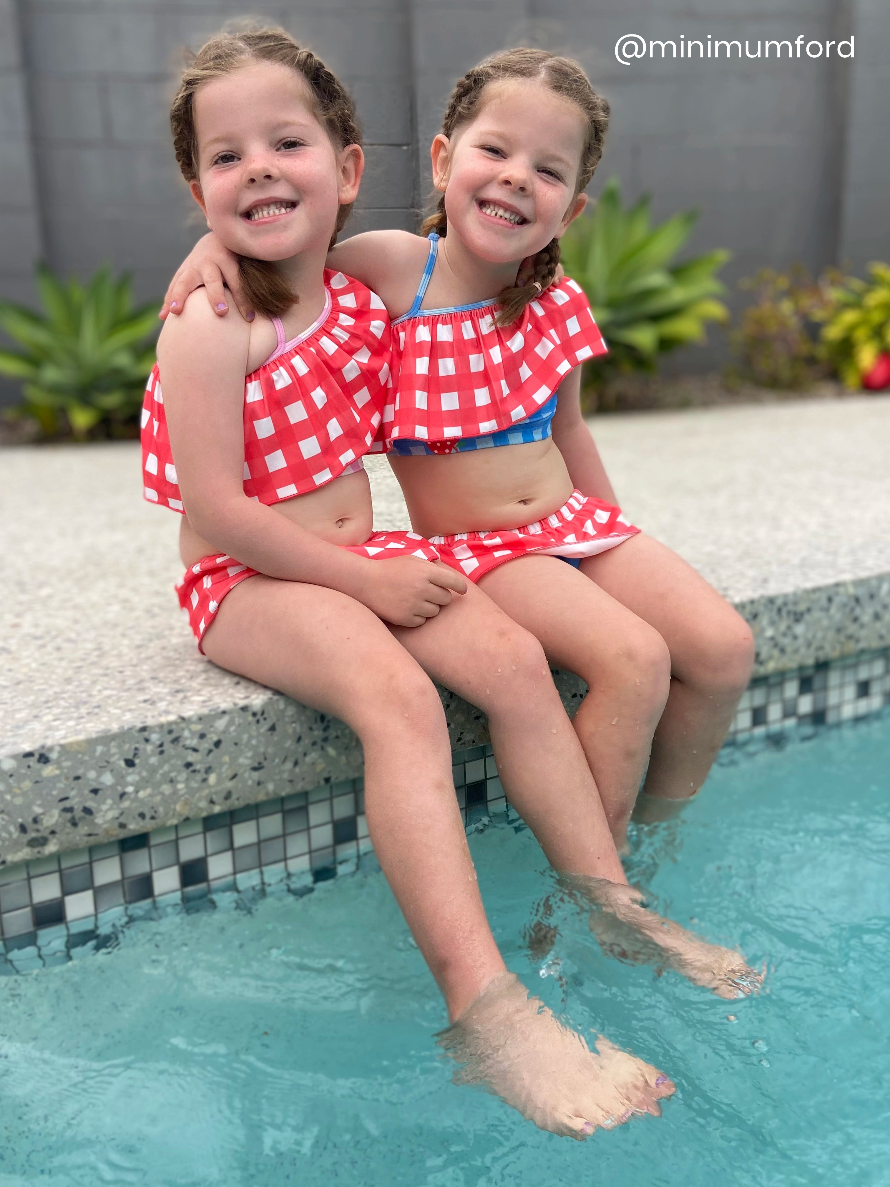 Two young girls in matching red and white checkered swimsuits sitting by a pool.