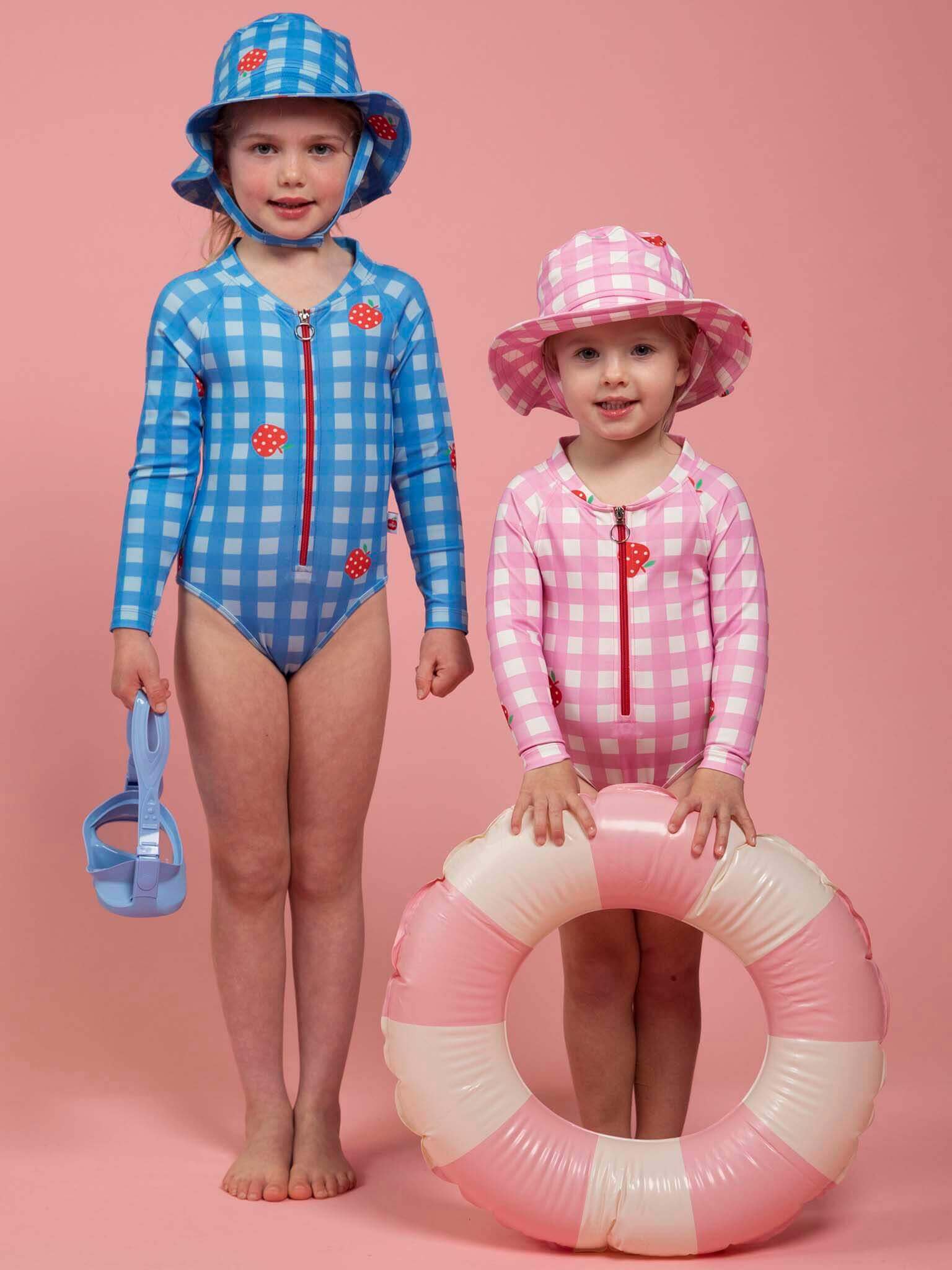 Two young children in Pink Gingham long-sleeve swimsuits and SPF 50 Gingham Swim Ponytail Hats stand against a pink background; one holds blue sandals, the other carries a pink and white swim ring.