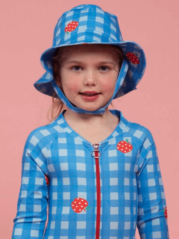 A young girl in a blue and white checkered outfit wears the Blue Gingham SPF 50 Gingham Swim Ponytail Hat, standing in front of a pink background.