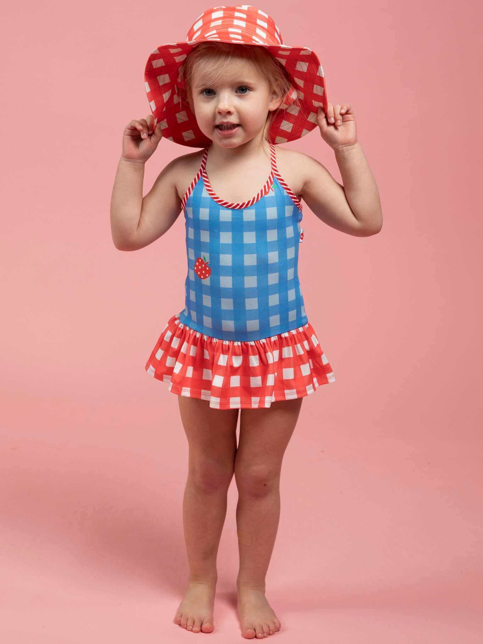 A young child stands on a pink background wearing a blue and red checkered swimsuit with the Red Gingham SPF 50 Gingham Swim Ponytail Hat for extra sun protection.