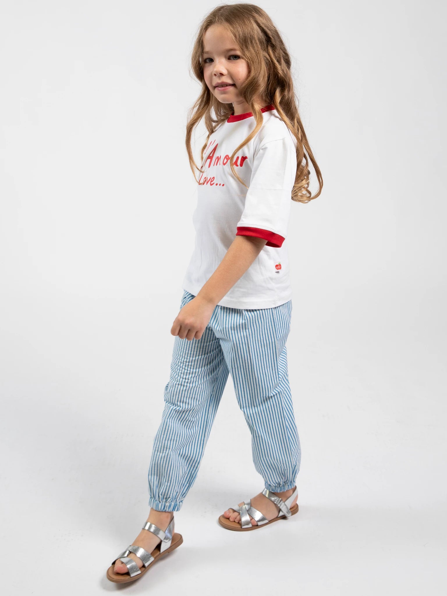 A young girl with long hair stands against a plain background, wearing Blue Stripe Balloon Pants from Blue Stripe, paired with a white tee with red trim from our girls collection and silver sandals.
