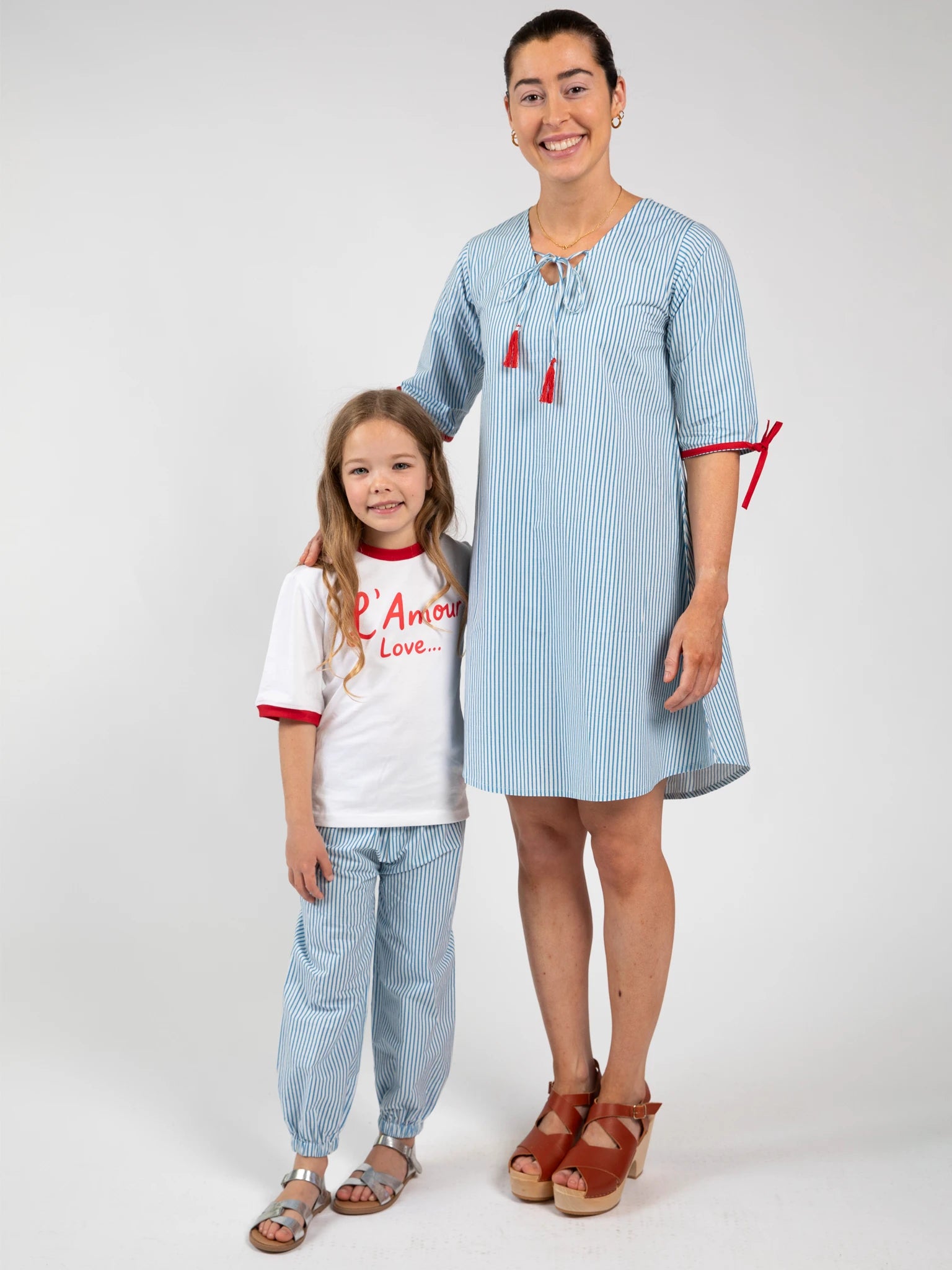 An adult and a child stand together. The adult wears a boho dress with blue and white stripes, red accents, and sandals, while the child wears Blue Stripe Balloon Pants by Blue Stripe and a white T-shirt with red text.