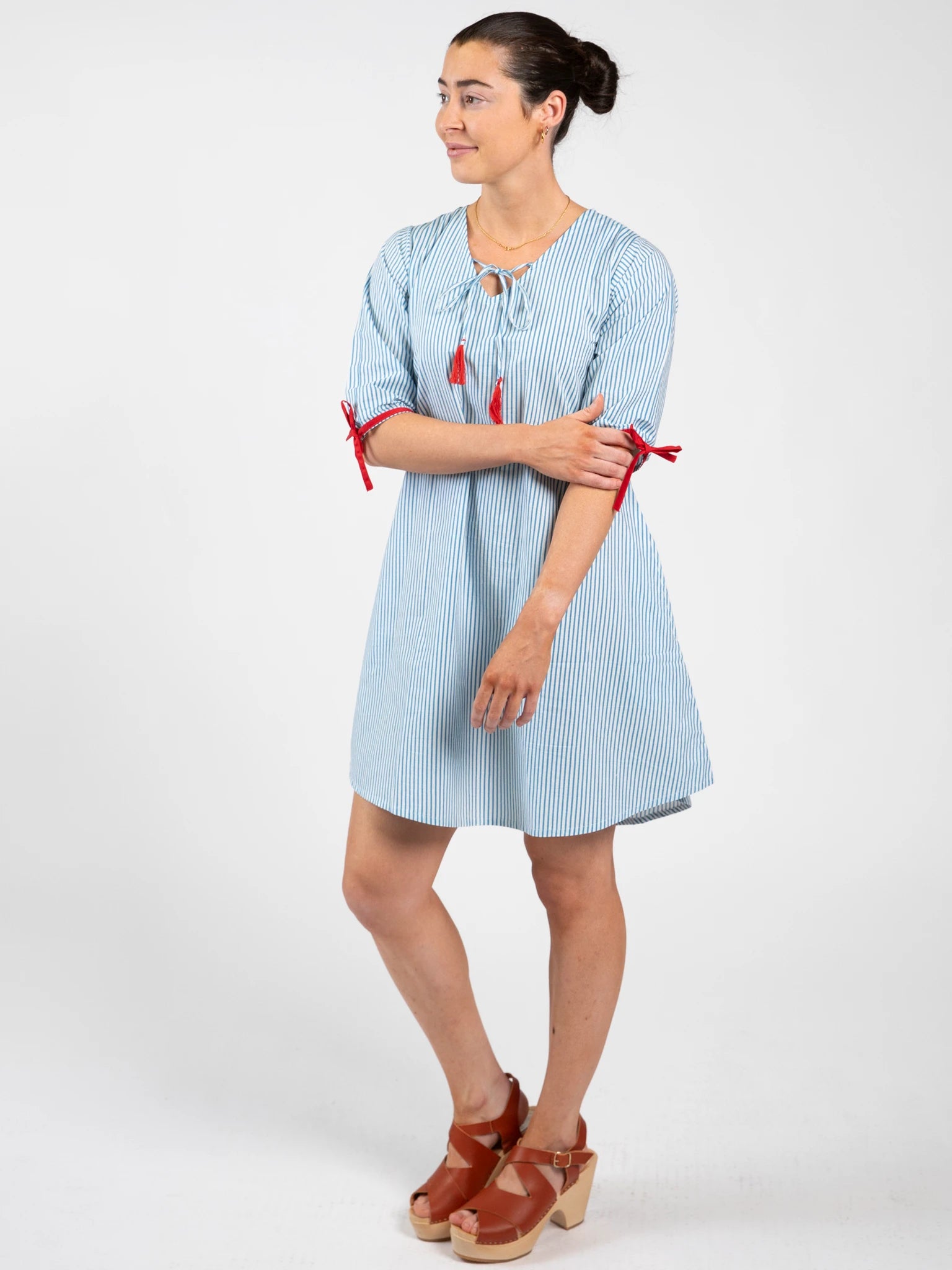 A woman wears the Blue Stripe Women's Harlow Kaftan Dress—100% cotton, blue and white stripes with red ribbon accents—by Blue Stripe, paired with brown sandals against a plain white background.