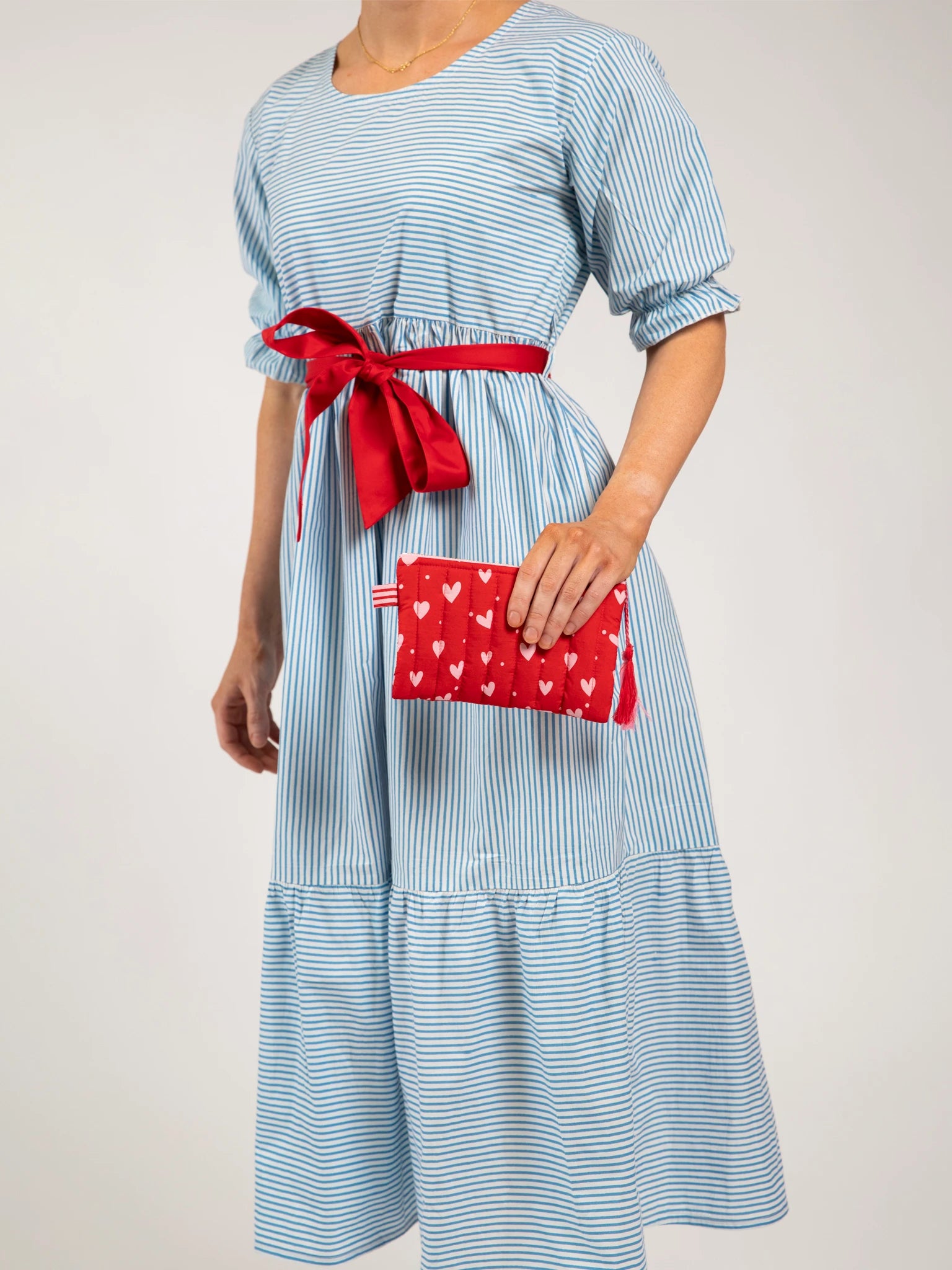 A person wearing the Blue Stripe Women's Blue Stripe Maxi Dress, styled with a red sash belt and holding a red clutch decorated with white hearts.