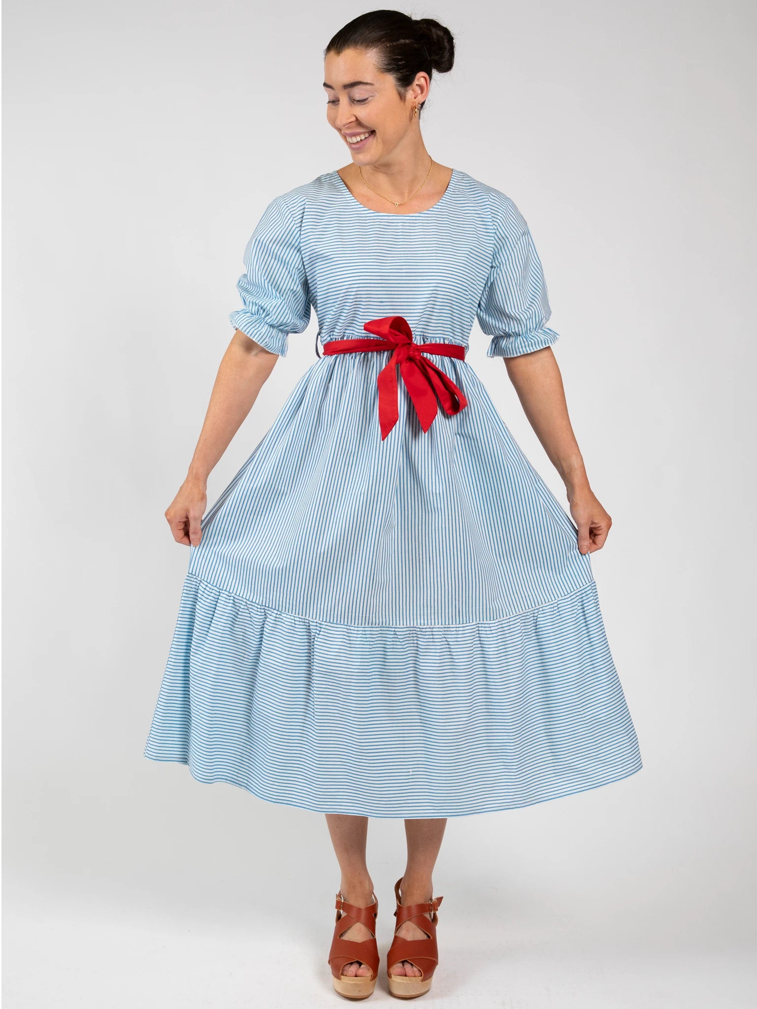 A woman stands against a plain background in the Blue Stripe Women's Blue Stripe Maxi Dress, featuring blue and white stripes and a red ribbon belt, paired with brown sandals for an effortlessly stylish, ethically made look.