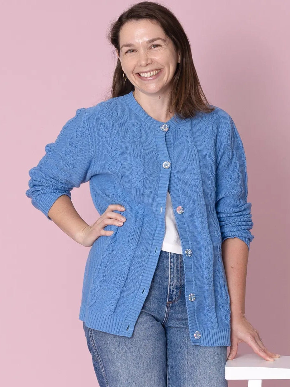 Woman wearing a blue cardigan and jeans standing next to a white stool on a pink background