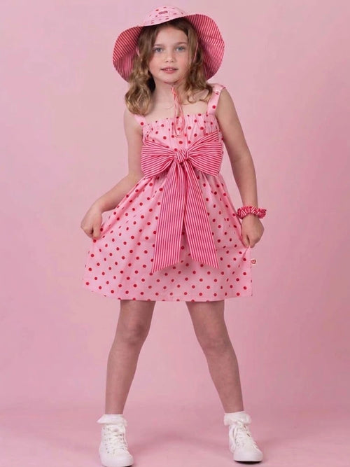 A young girl modeling a pink and white polka dot dress with a striped bow at the waist, a matching pink and white polka dot sun hat, and white sneakers, set against a solid pink studio background.