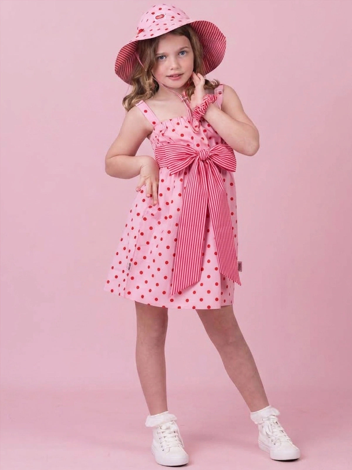 A young girl modeling a pink and white striped sun hat, a matching pink polka-dot dress with a large bow, and white sneakers, set against a solid pink studio background.
