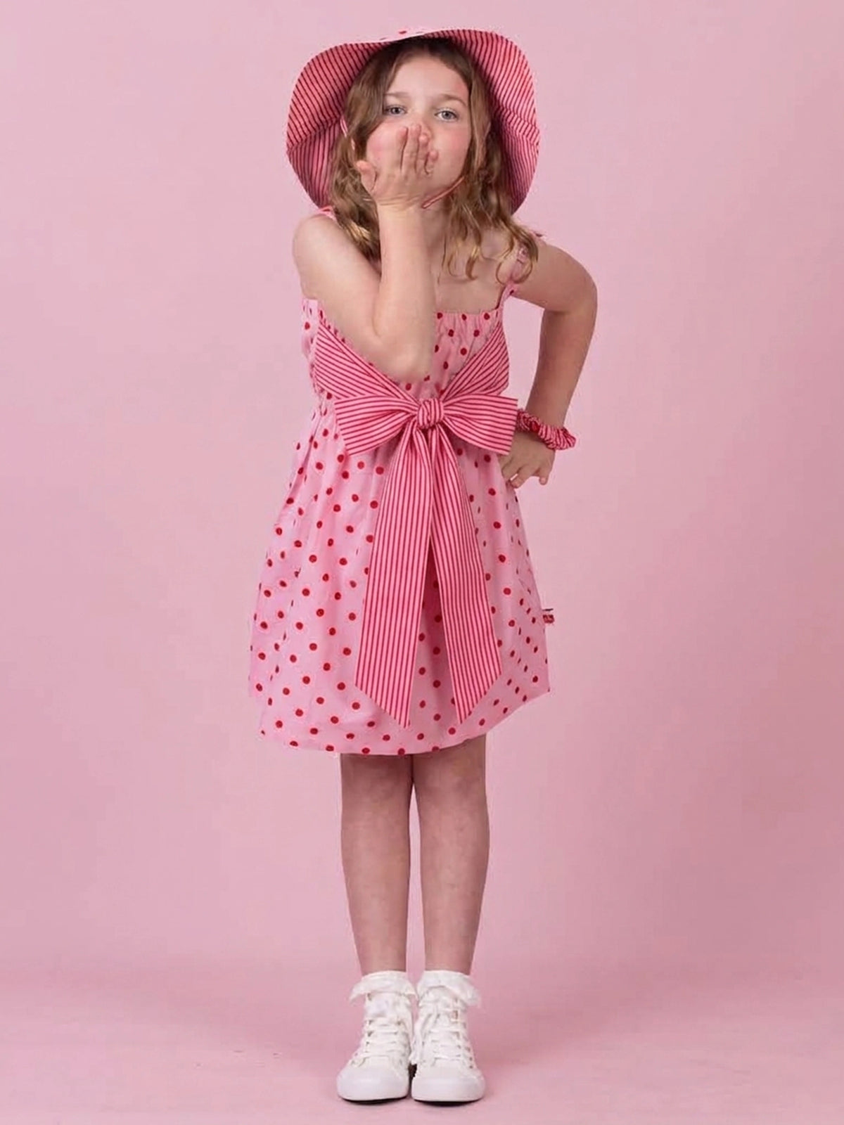 A young girl wearing a pink and white striped dress with red polka dots, a matching striped sun hat, and white sneakers, posing against a solid pink background.