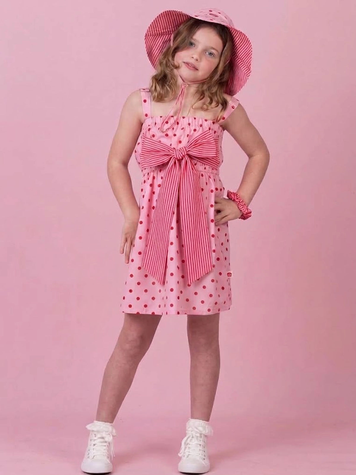 A young girl models a pink and white striped dress with red polka dots, featuring a large matching bow at the waist, a striped sun hat, and white sneakers, set against a solid pink background.