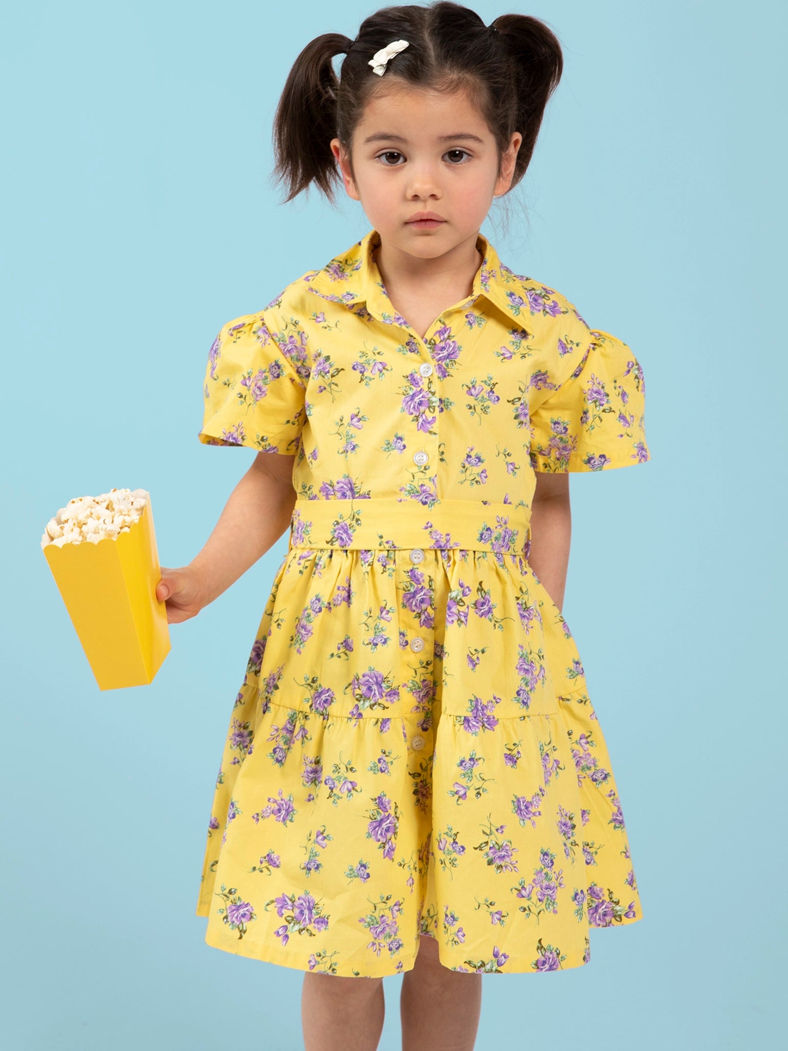 Young girl wearing a yellow floral Buttercup dress holding a yellow popcorn bucket against a light blue background