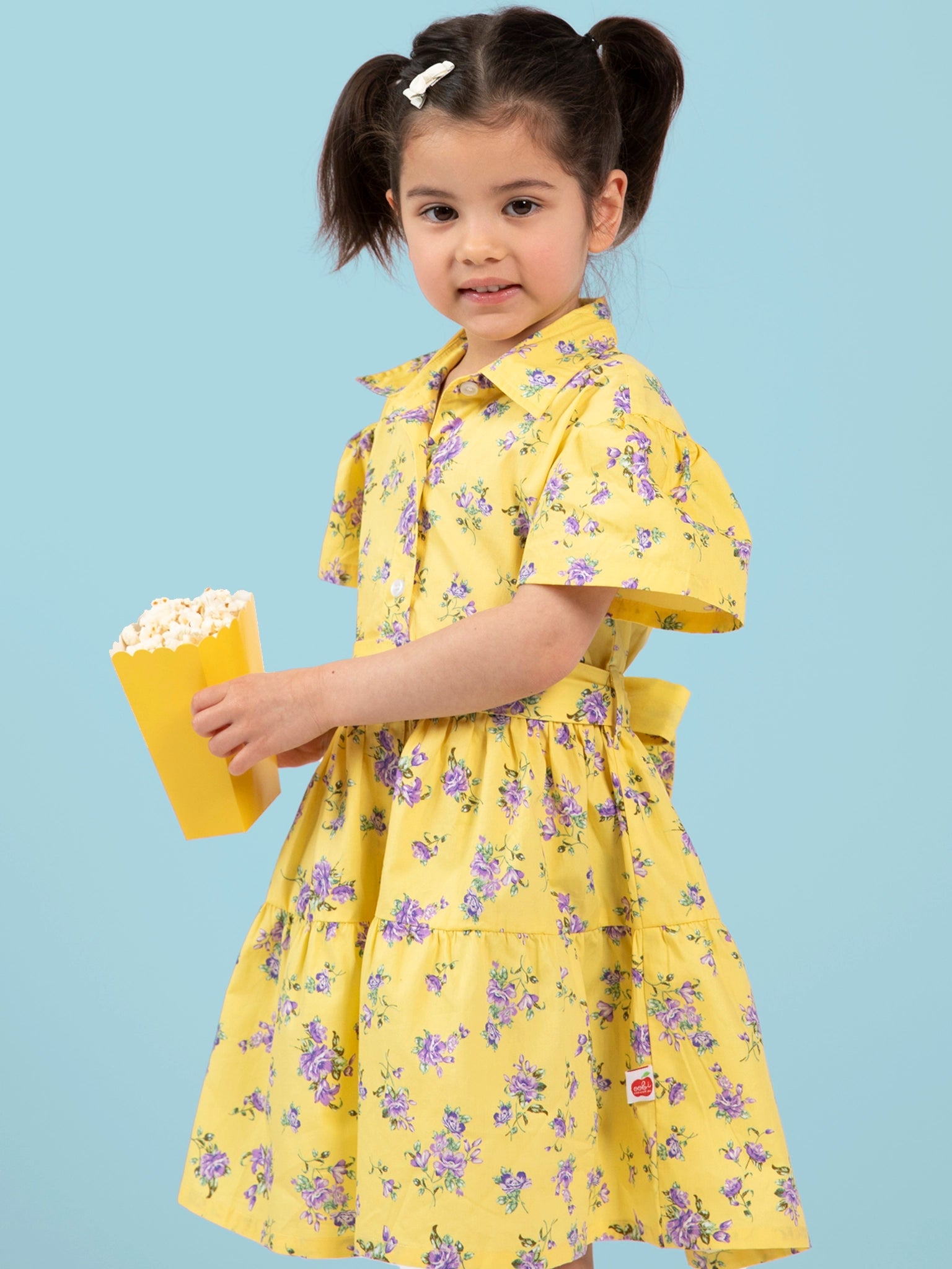 Child wearing a yellow floral Buttercup dress holding popcorn against a light blue background