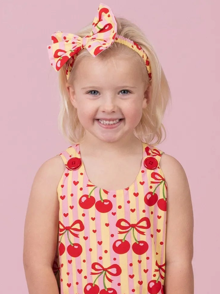 A young girl with blonde hair wearing a pink and red cherry print sleeveless top and a matching cherry print bow headband, set against a solid pink background.