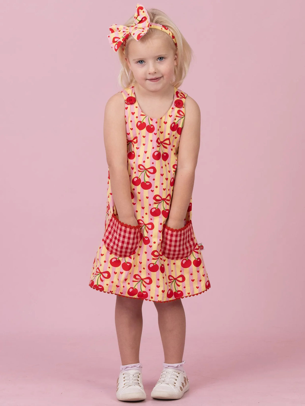 A young girl stands against a solid pink studio background wearing a sleeveless cream dress with a red cherry print, red gingham pocket trim, and a matching red-and-cream bow headband.
