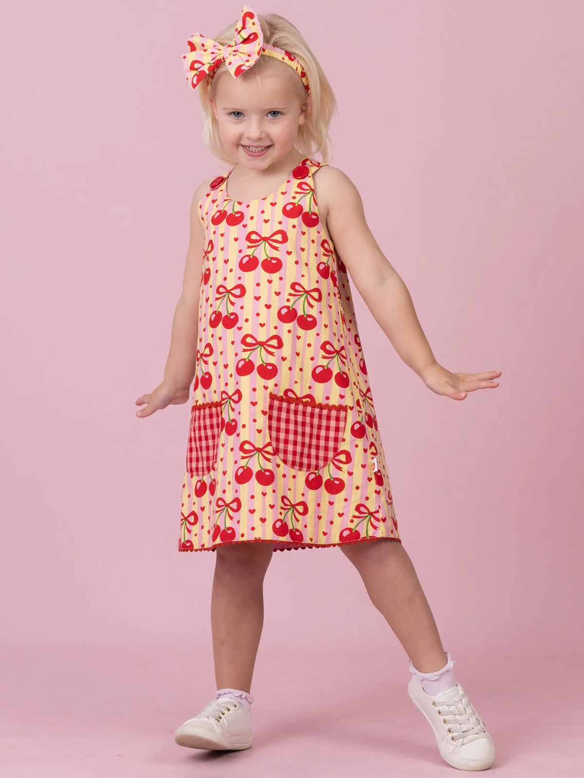 A young girl modeling a sleeveless dress with a red cherry and bow pattern on a cream background, featuring red gingham pockets, a matching red and cream bow headband, and white sneakers, set against a solid pink studio background.