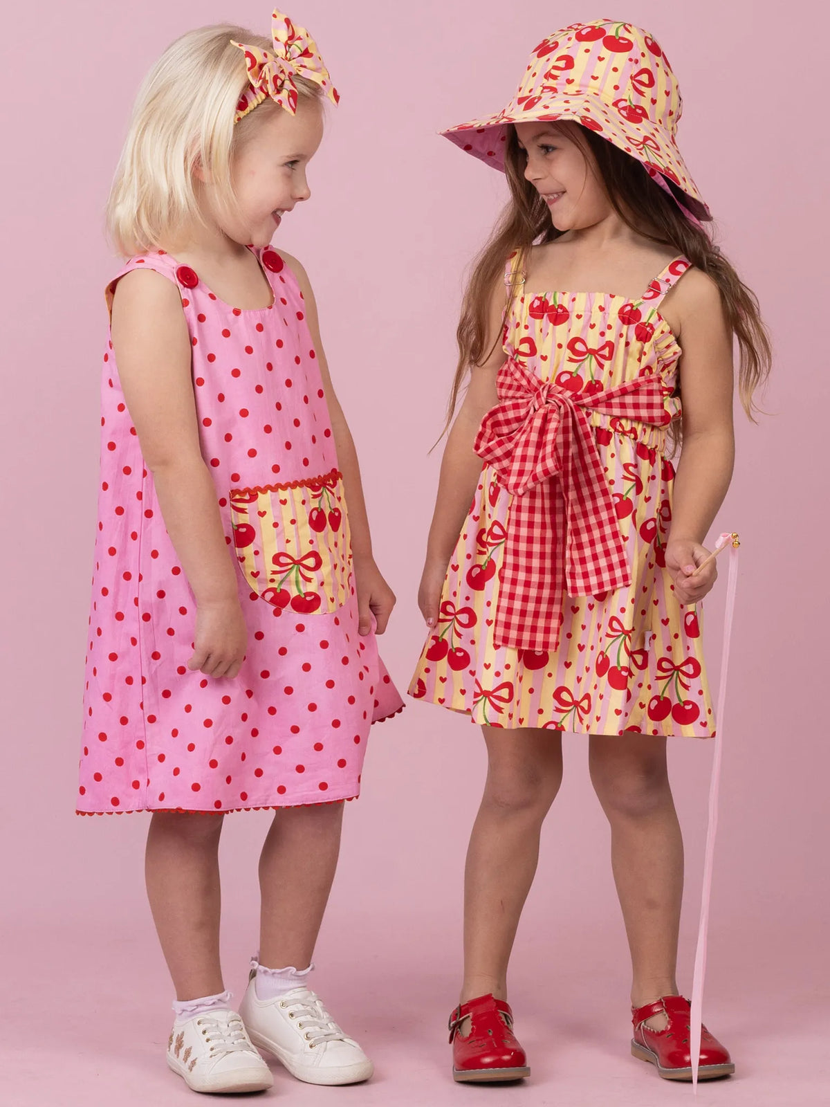 Two girls in colourful dresses. The girl on the left wears a pink polka dot dress and hair bow and the girl on the right wears a cherry print dress and hat.