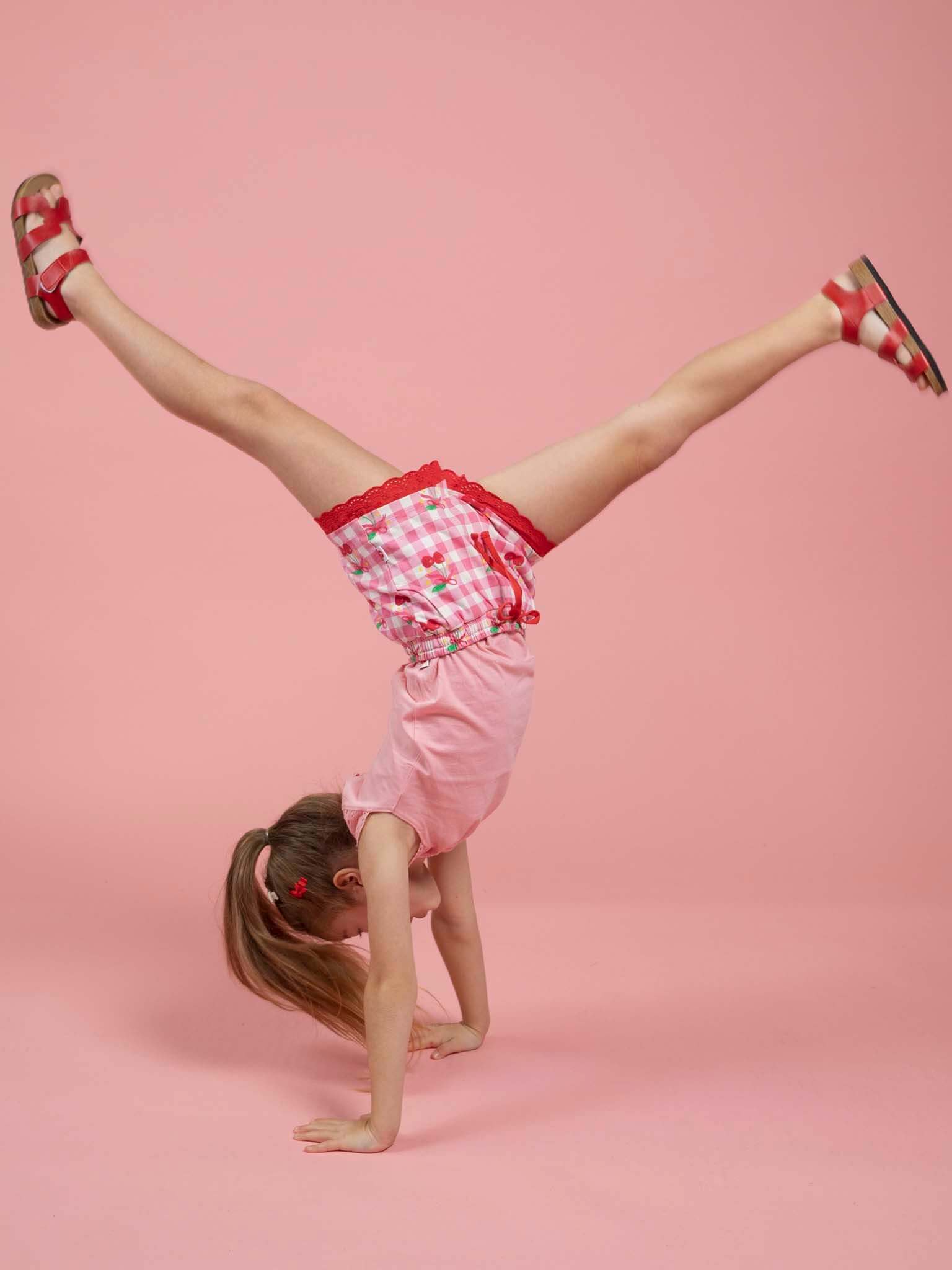 A young child wearing Cherry Gingham Jersey Shorts by Cherry Gingham performs a handstand against a solid pink background.