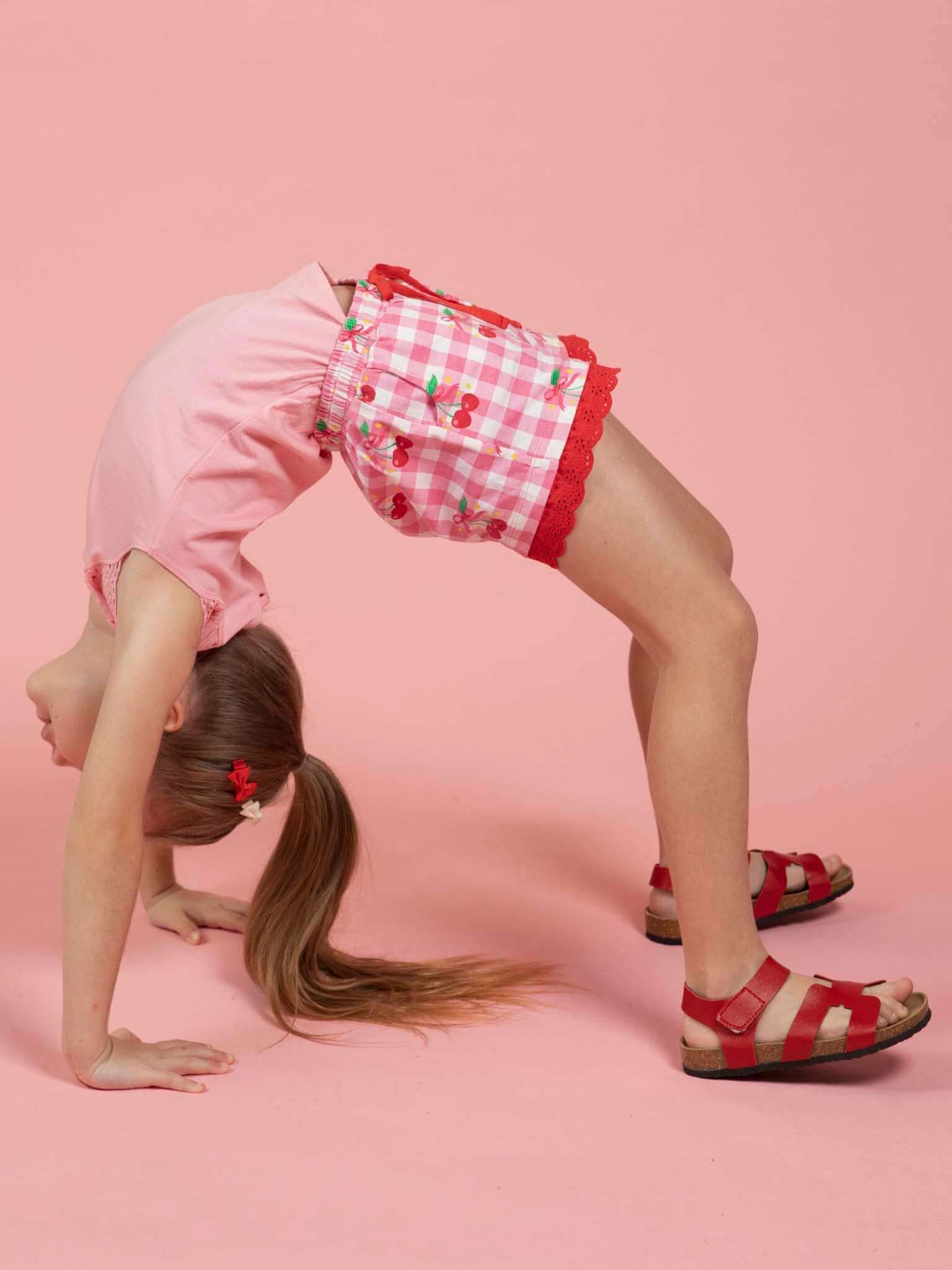 A young girl with a ponytail does a backbend on a pink background, wearing a pink shirt, Cherry Gingham Jersey Shorts by Cherry Gingham, and red sandals.
