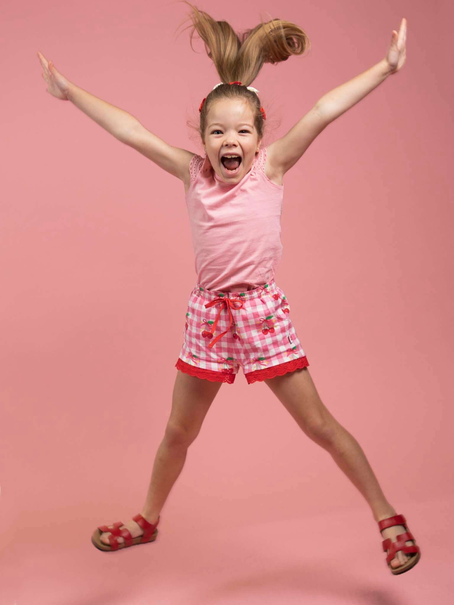 A young girl with brown hair jumps in the air, wearing a pink shirt, Cherry Gingham Jersey Shorts by Cherry Gingham, and red sandals against a pink background.