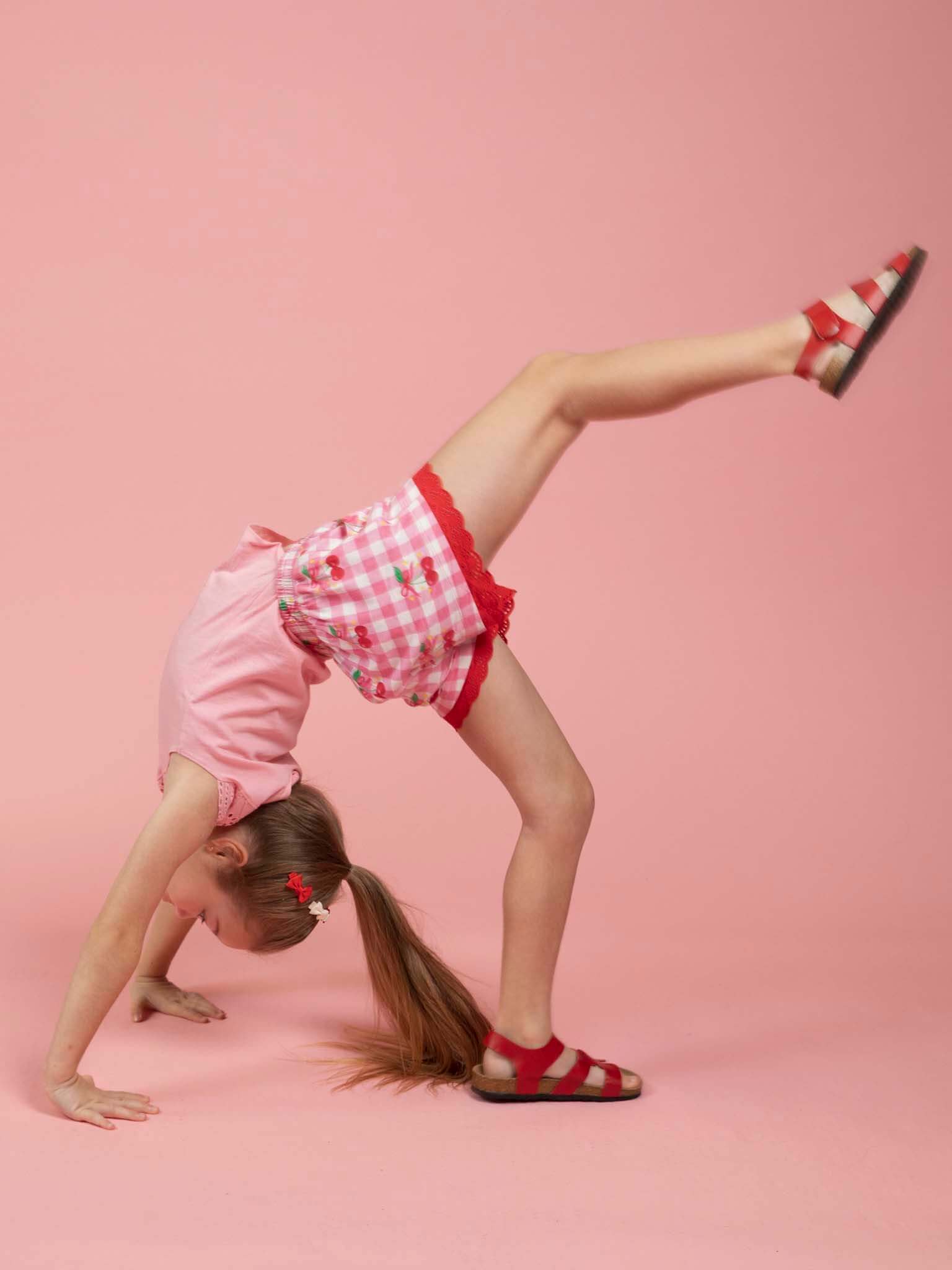 A young girl in Cherry Gingham Jersey Shorts and red sandals performs a cartwheel on a pink background, her Cherry Gingham shorts adding comfort and style to her playful look.