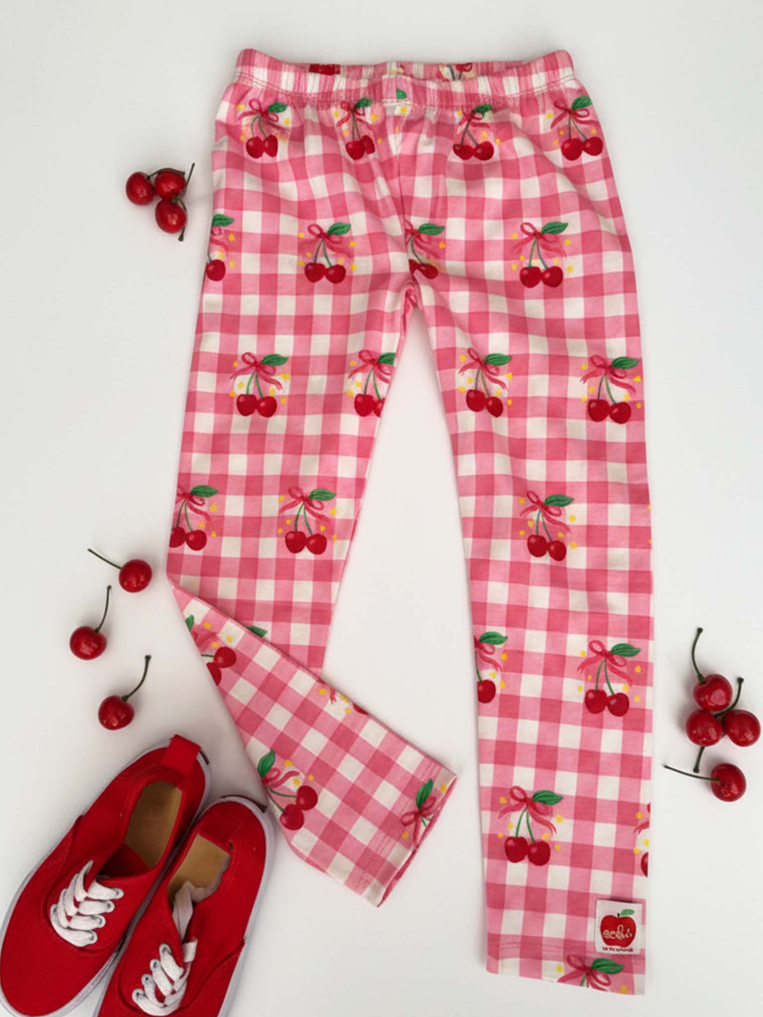 Cherry Gingham Jersey Leggings by Cherry Gingham in pink and white gingham with a cherry print are laid flat, surrounded by fresh cherries and red sneakers on a white background.