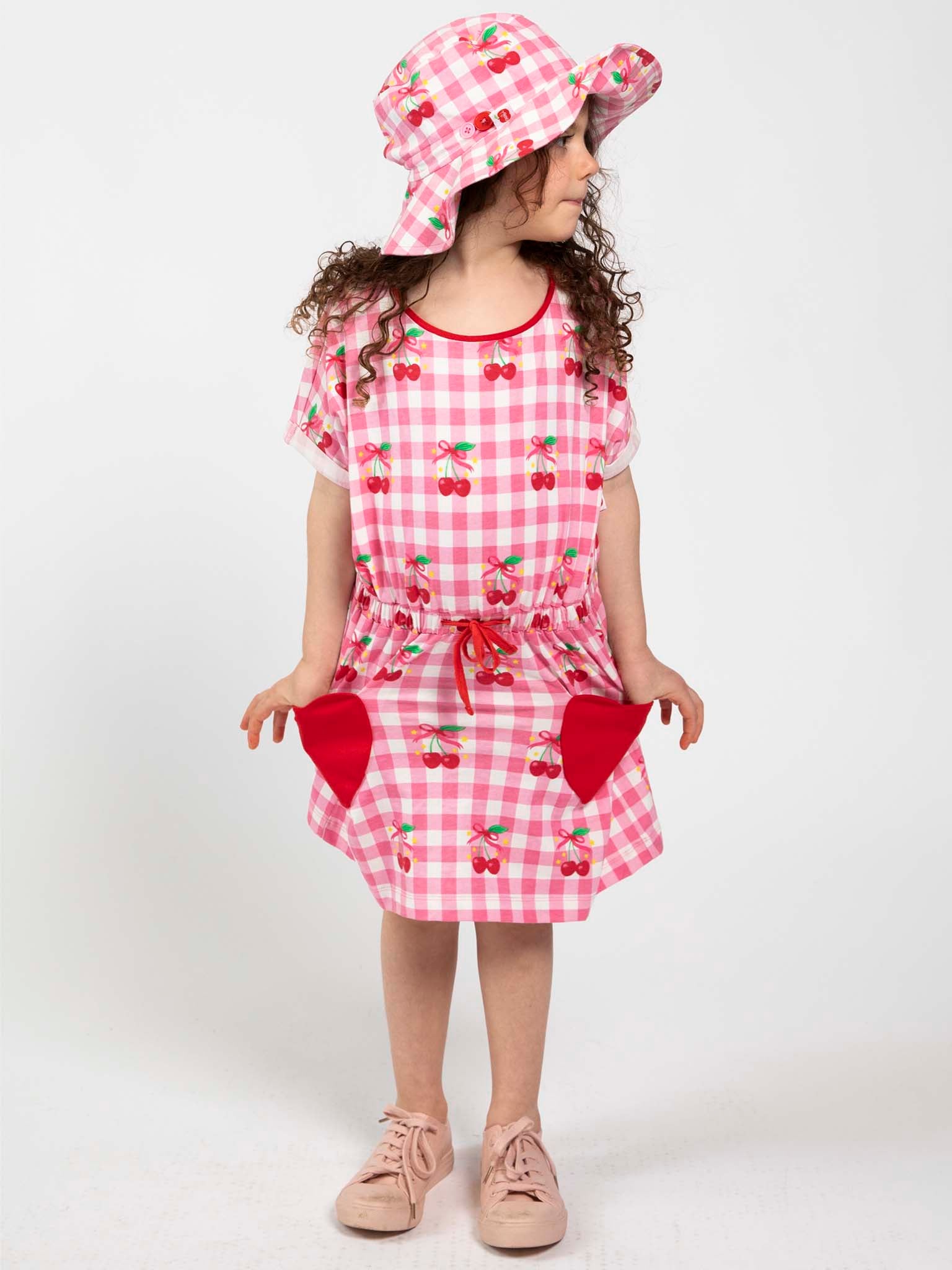 A young child wears the Cherry Gingham Ponytail Hat and a matching pink and white checkered dress with cherry prints and red pockets from Cherry Gingham, standing against a plain white background.