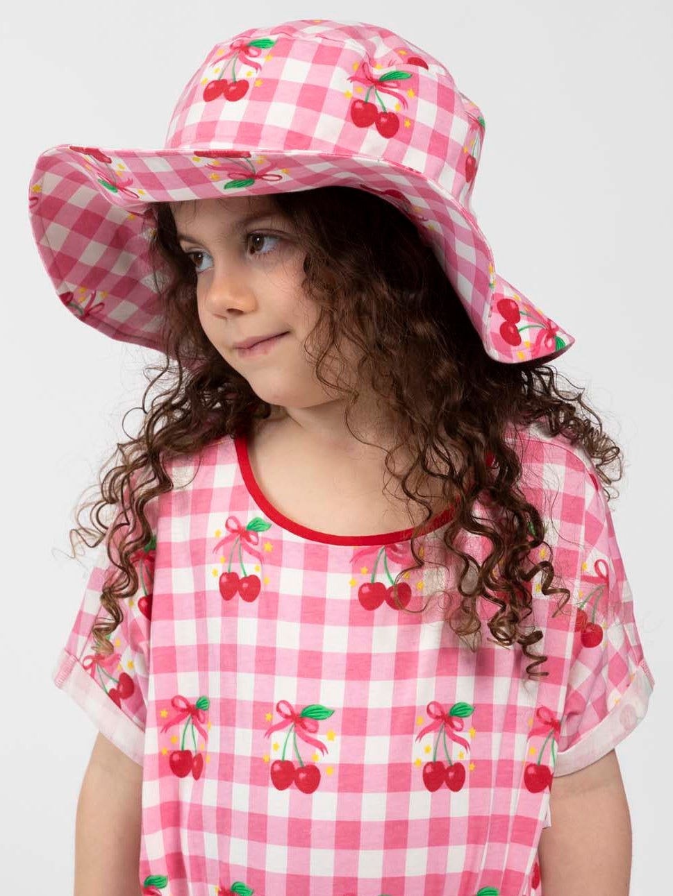 Child wearing a pink gingham dress with cherry pattern and matching hat on a white background