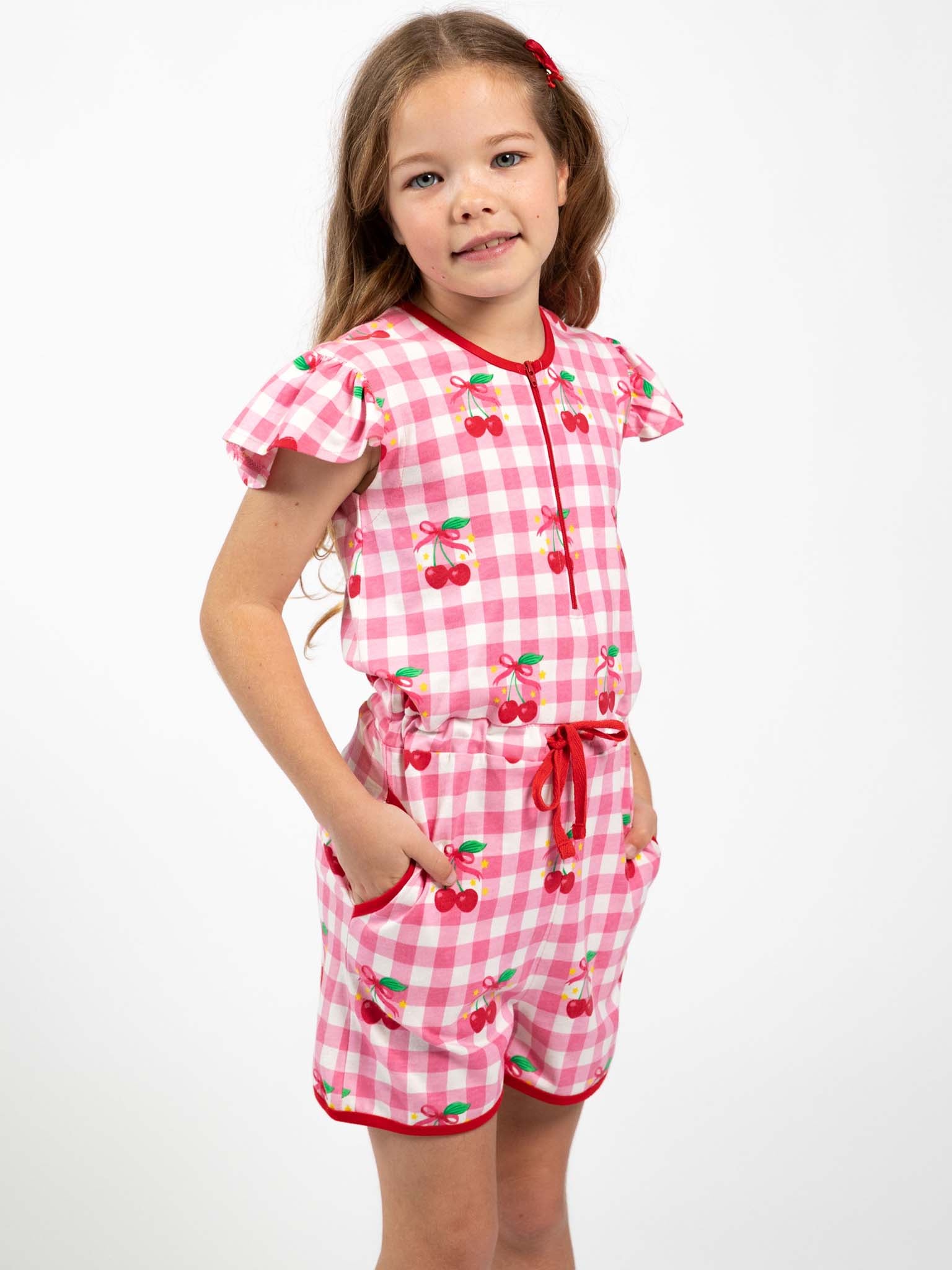 Young girl wears the Cherry Gingham Jersey Playsuit by Cherry Gingham, featuring a cherry print and red trim, standing against a plain white background.