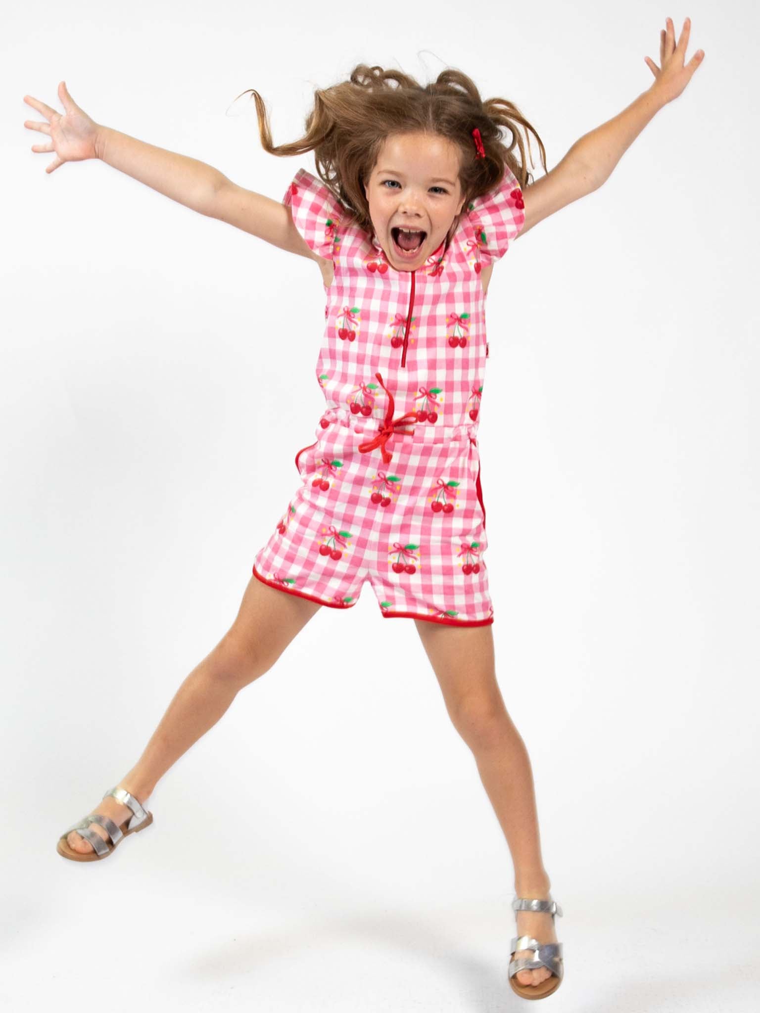 A young girl in a Cherry Gingham Jersey Playsuit by Cherry Gingham jumps excitedly in sandals against a plain white background.