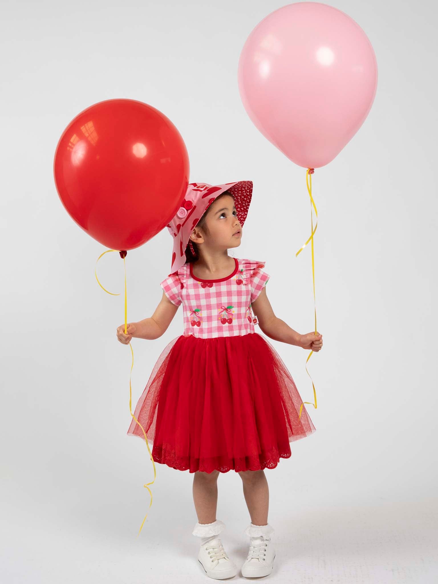 A young girl wears the Cherry Gingham Tutu Dress by Cherry Gingham, holding a red balloon in one hand and a pink balloon in the other, standing against a plain background.