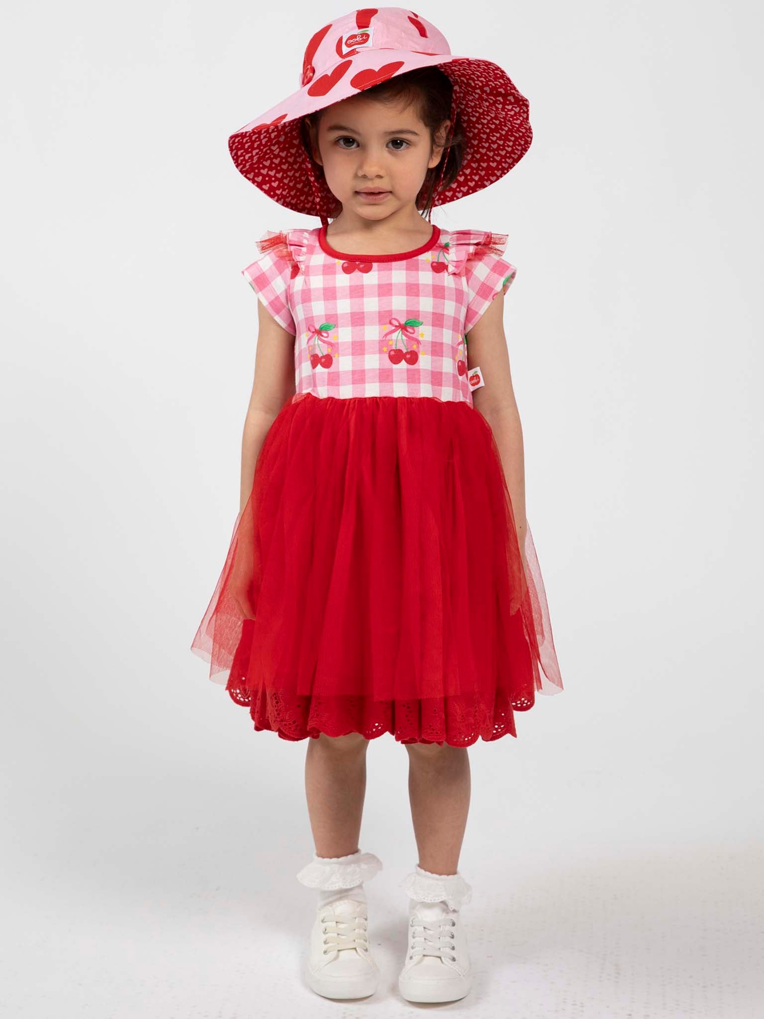 A young girl faces forward, wearing the Cherry Gingham Tutu Dress by Cherry Gingham with a red tulle skirt, white shoes, frilly socks, and a wide-brimmed hat in matching red and pink patterns.