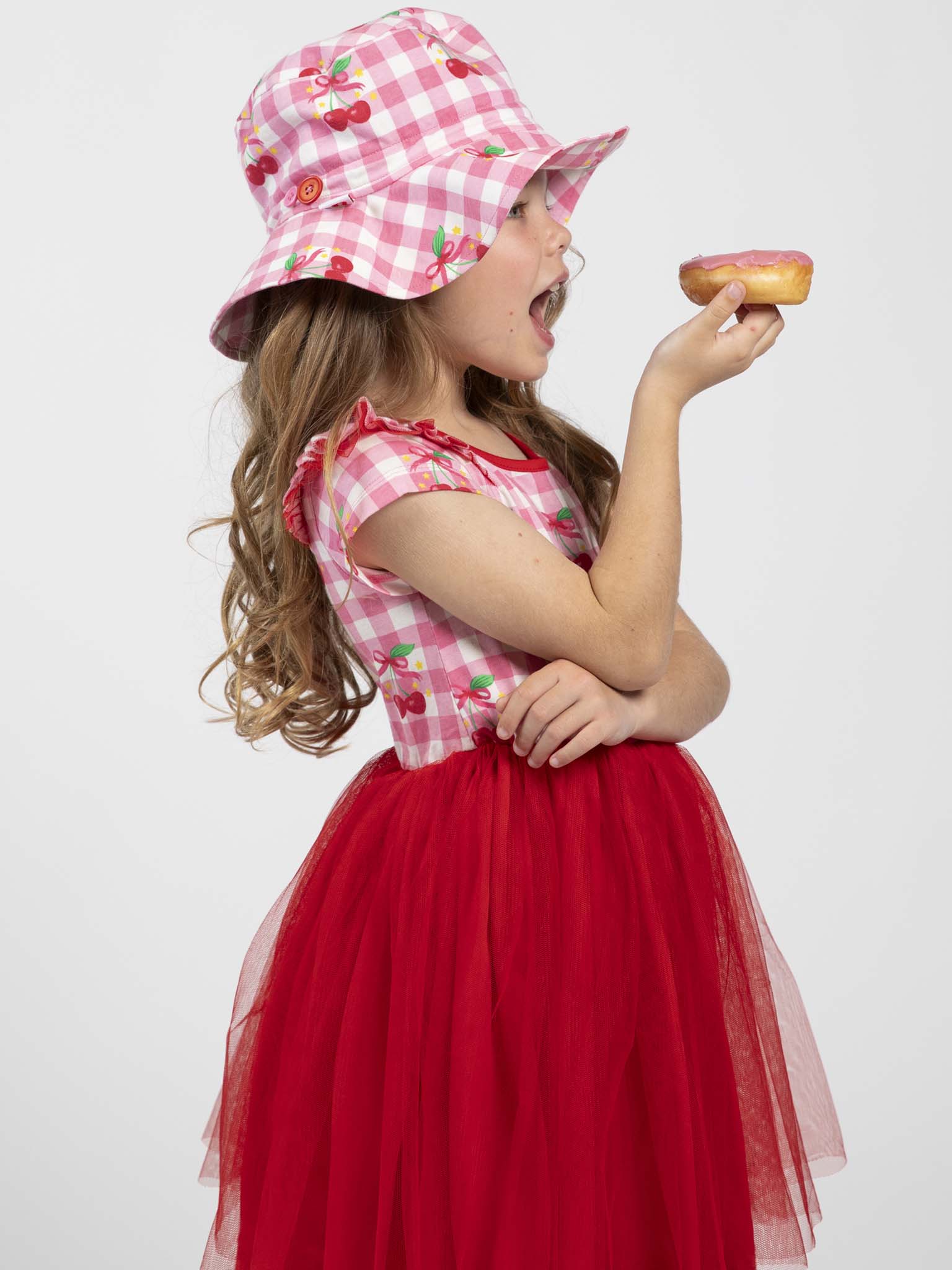 A young girl, wearing the Cherry Gingham Tutu Dress and matching hat by Cherry Gingham, holds a donut and looks ready to take a bite while standing against a plain background.