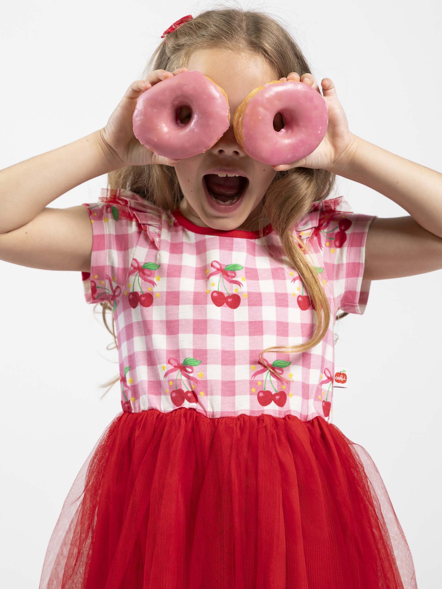 A young girl in the Cherry Gingham Tutu Dress by Cherry Gingham holds two pink-frosted donuts over her eyes, her mouth open in excitement.