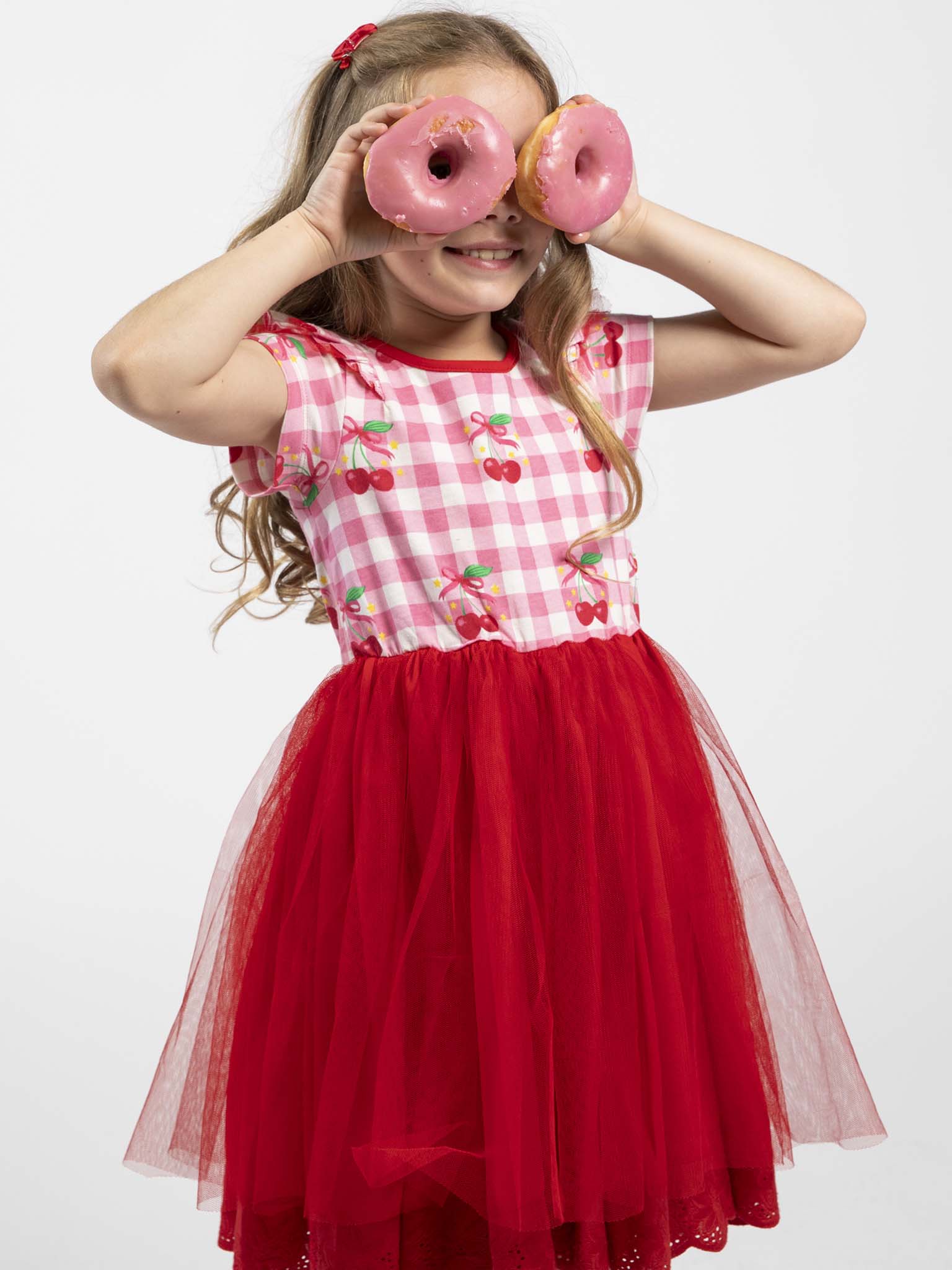 A young girl in a Cherry Gingham Tutu Dress by Cherry Gingham playfully holds two pink frosted donuts over her eyes, smiling against a plain white background.