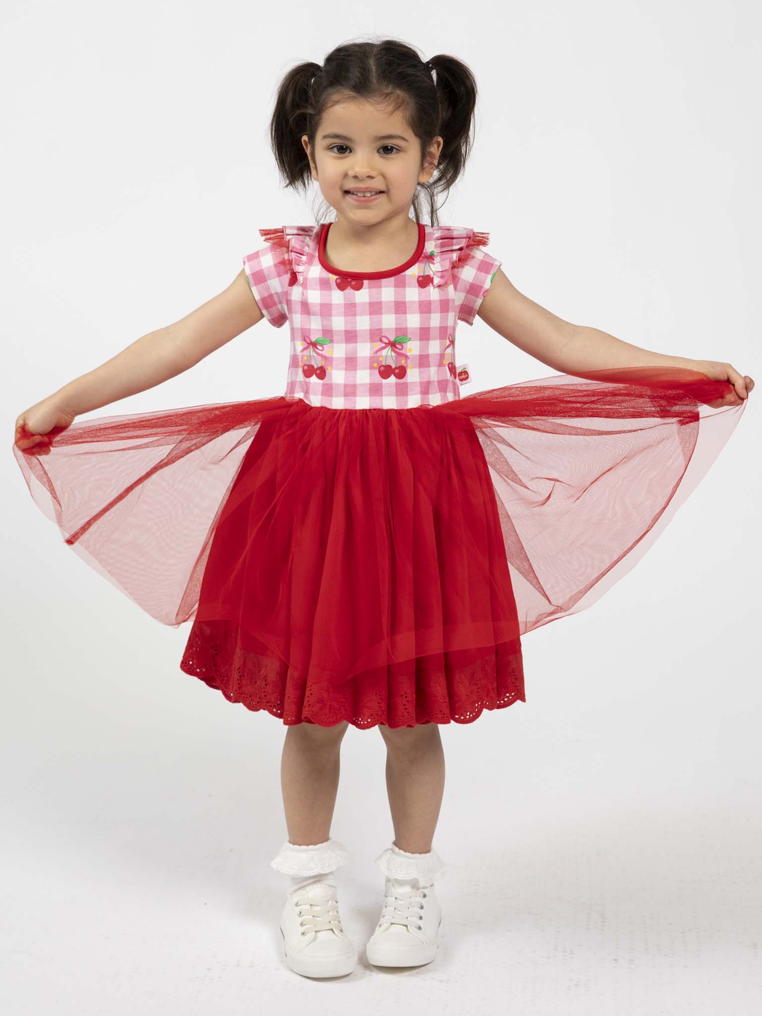 A young girl smiles while wearing the Cherry Gingham Tutu Dress by Cherry Gingham, featuring hand-printed cherry patterns and gingham design, paired with white socks and sneakers on a plain background.