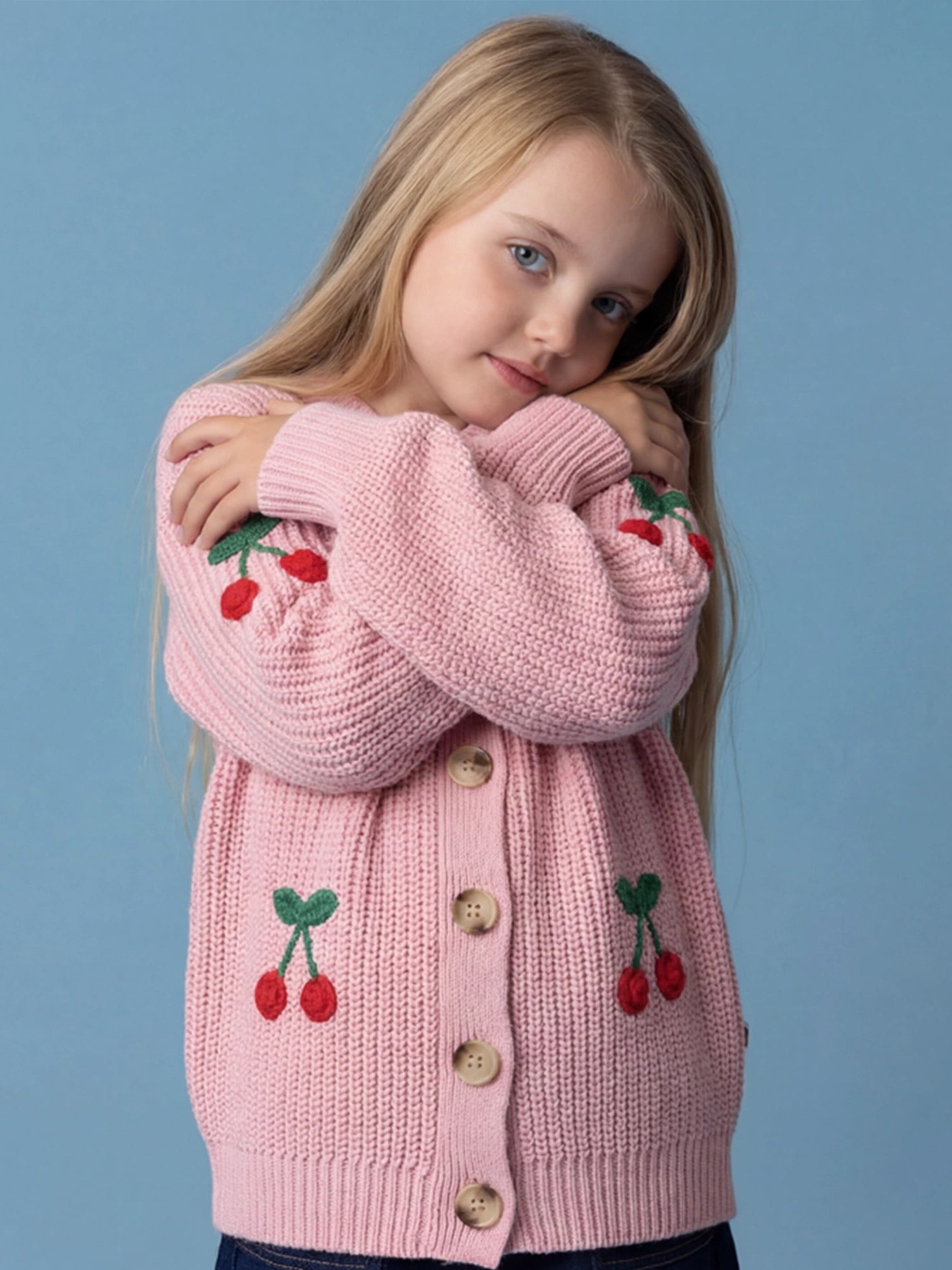A young girl with long blonde hair models the Knitwear Cherry Appliqué Cardigan, featuring a chunky pink knit and cherry details, standing against a plain blue background with her arms crossed.