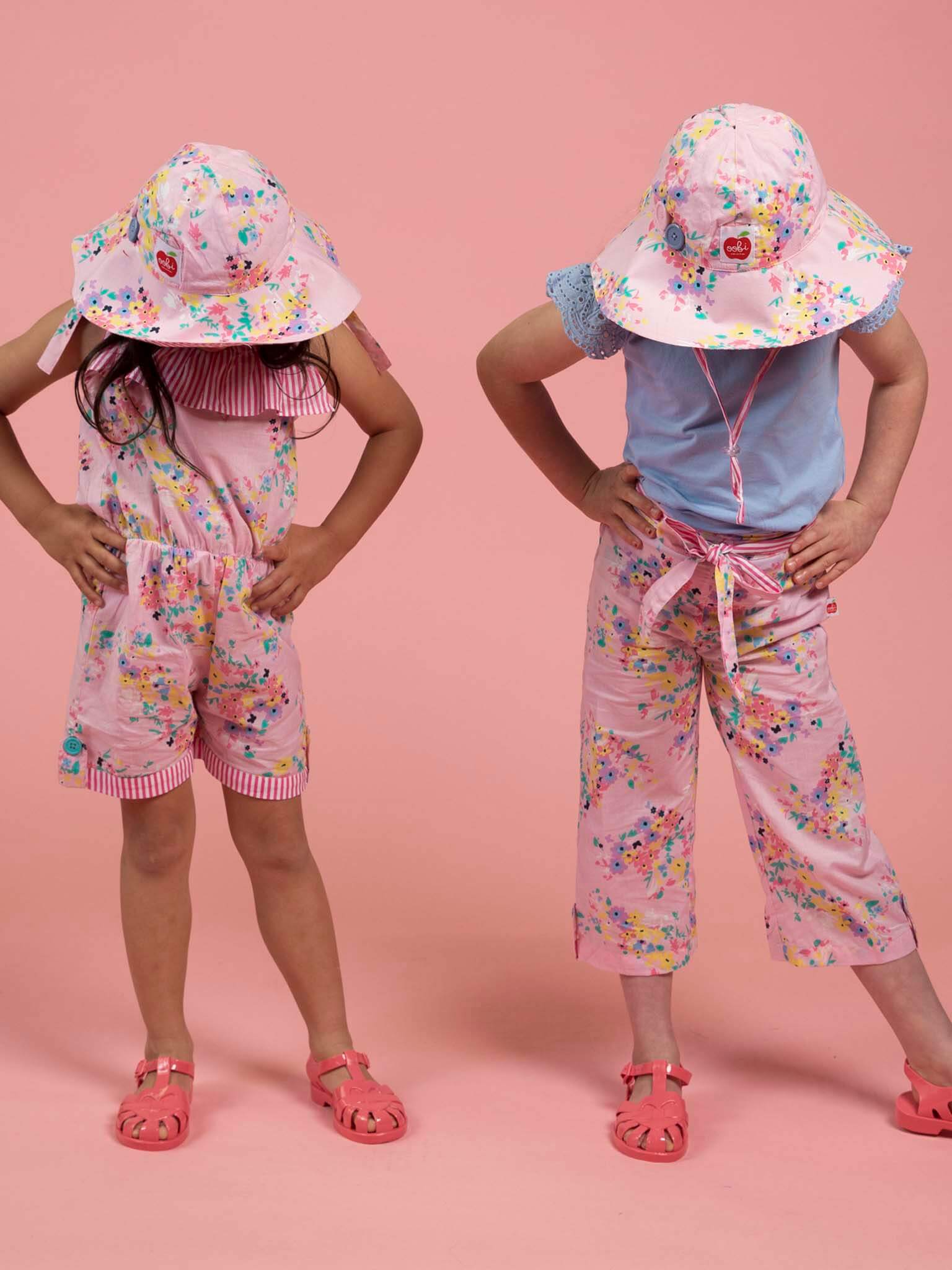 Two children wearing Confetti Floral Ponytail Hats and matching floral pink outfits stand with hands on hips against a pink background, looking down.
