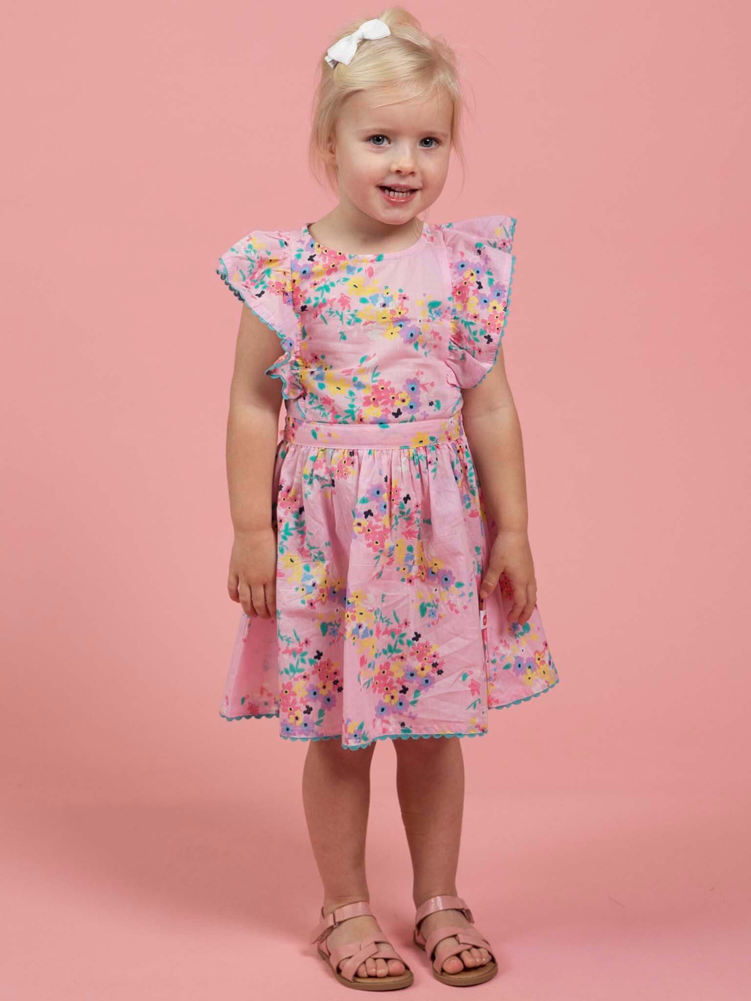 A young girl poses against a pink backdrop in the Confetti Floral Lacy Dress by Confetti Floral, styled with sandals, a white hair bow, and a summer hat for an irresistibly charming look.