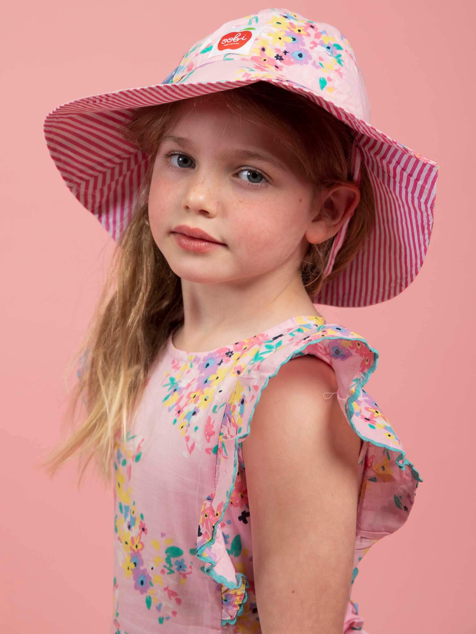 A young girl wears the Confetti Floral Lacy Dress by Confetti Floral and a wide-brimmed pink striped hat, standing against a solid pink background.