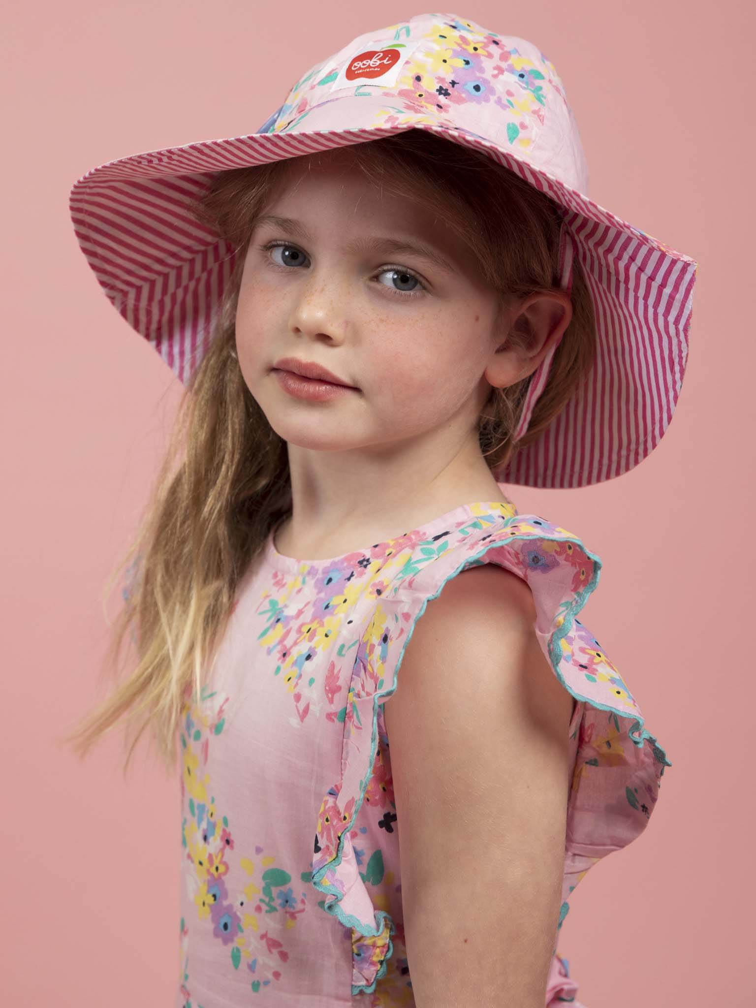 A young girl in the Confetti Floral Lacy Dress by Confetti Floral, paired with a wide-brimmed summer hat featuring stripes and a red logo, stands against a solid pink background.