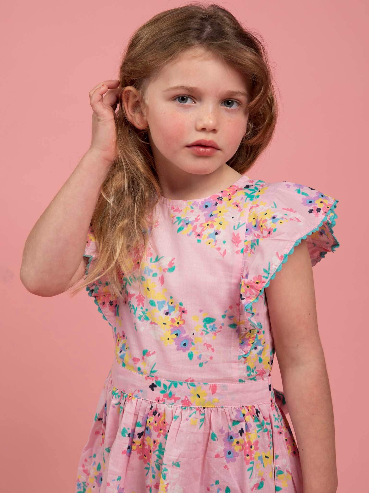 A young girl with long light brown hair wears the Confetti Floral Lacy Dress by Confetti Floral, featuring pink floral ruffle sleeves, standing against a solid pink background with one hand touching her ear.