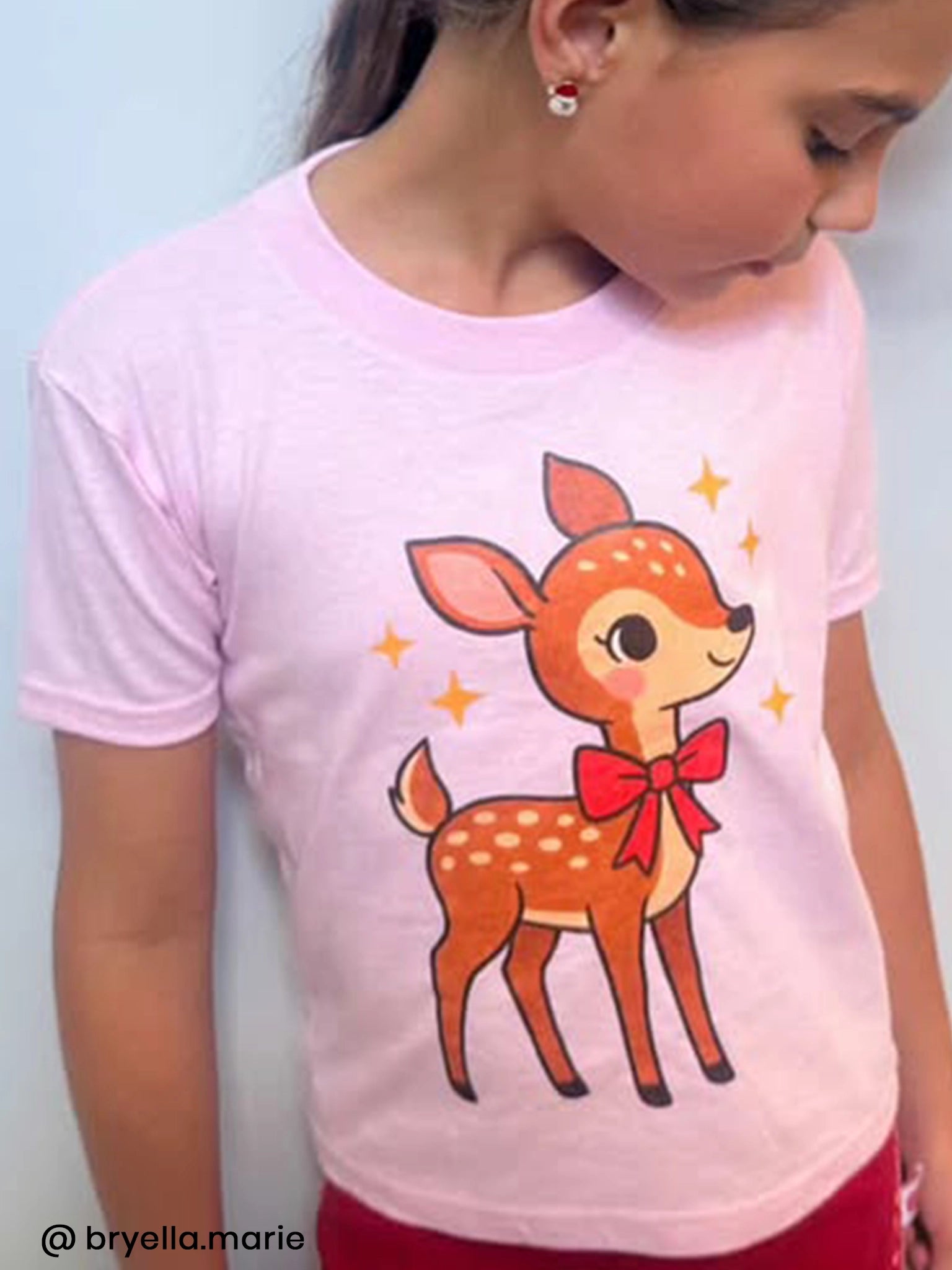 A girl wears the Essentials Deer Printed Tee—an oversized, light pink shirt with a cartoon fawn, red bow, and sparkles on the front. Personalisation available; sizes from 2Y to adult.