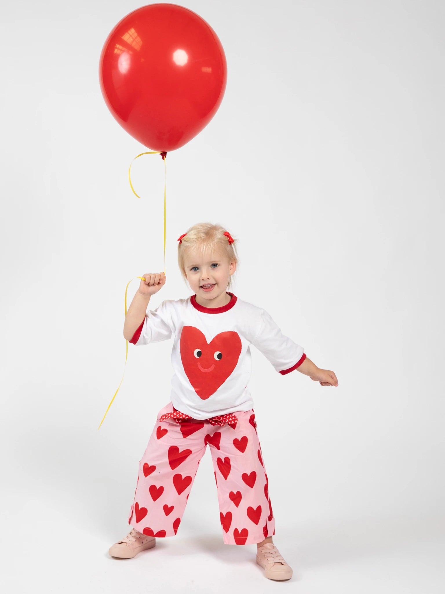 A young child models Big Hearts' Big Heart Wide-Leg Pants, featuring a hand-printed heart design and elastic waist, while holding a red balloon against a plain white background.