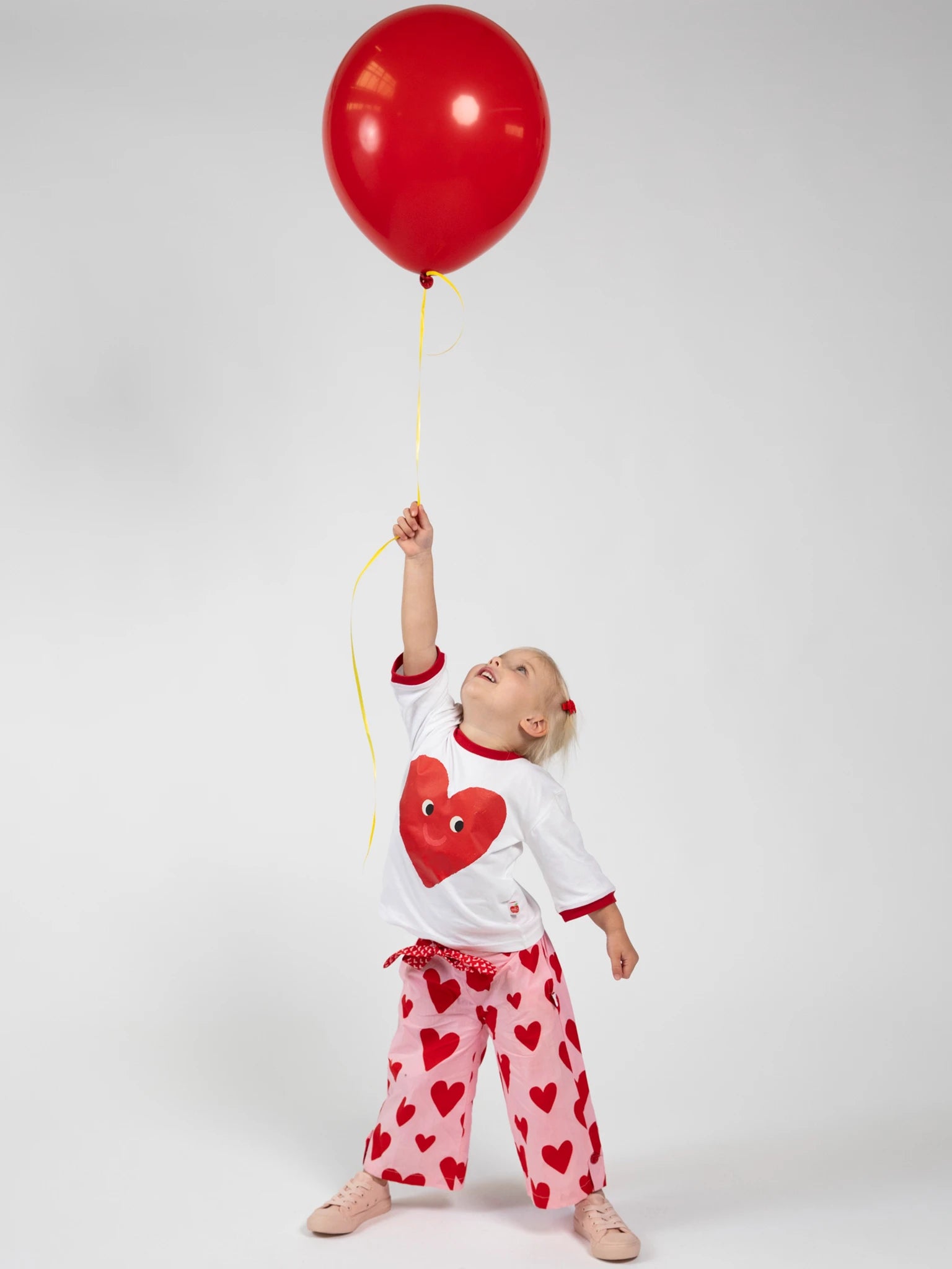 A young child in Big Hearts' Big Heart Wide-Leg Pants with an elastic waist holds a red balloon by its yellow string, set against a plain white background.