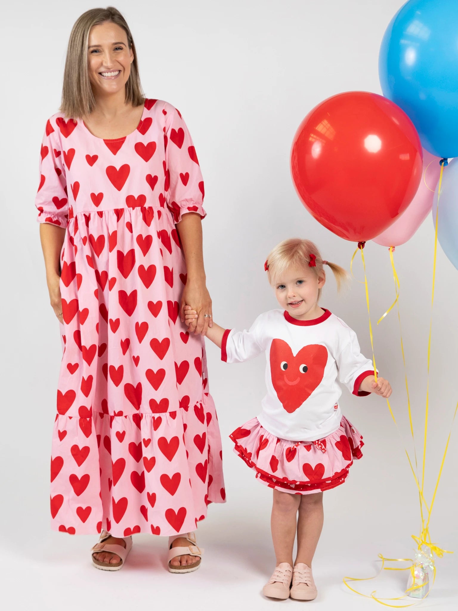 A woman and child hold hands in matching Big Hearts outfits. The woman wears the Women's Big Heart Maxi Dress, and the child holds red and blue balloons against a plain white background.