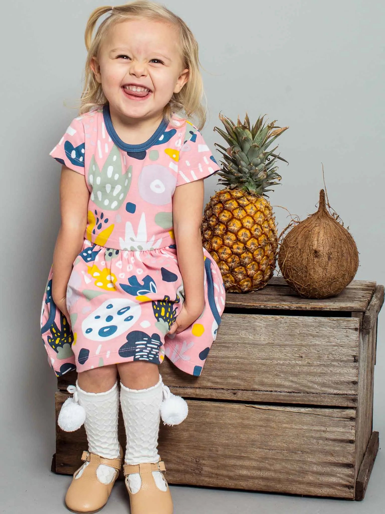 A sitting and smiling girl wearing a pink jungle print dress and white pom pom socks next to a pineapple and coconut