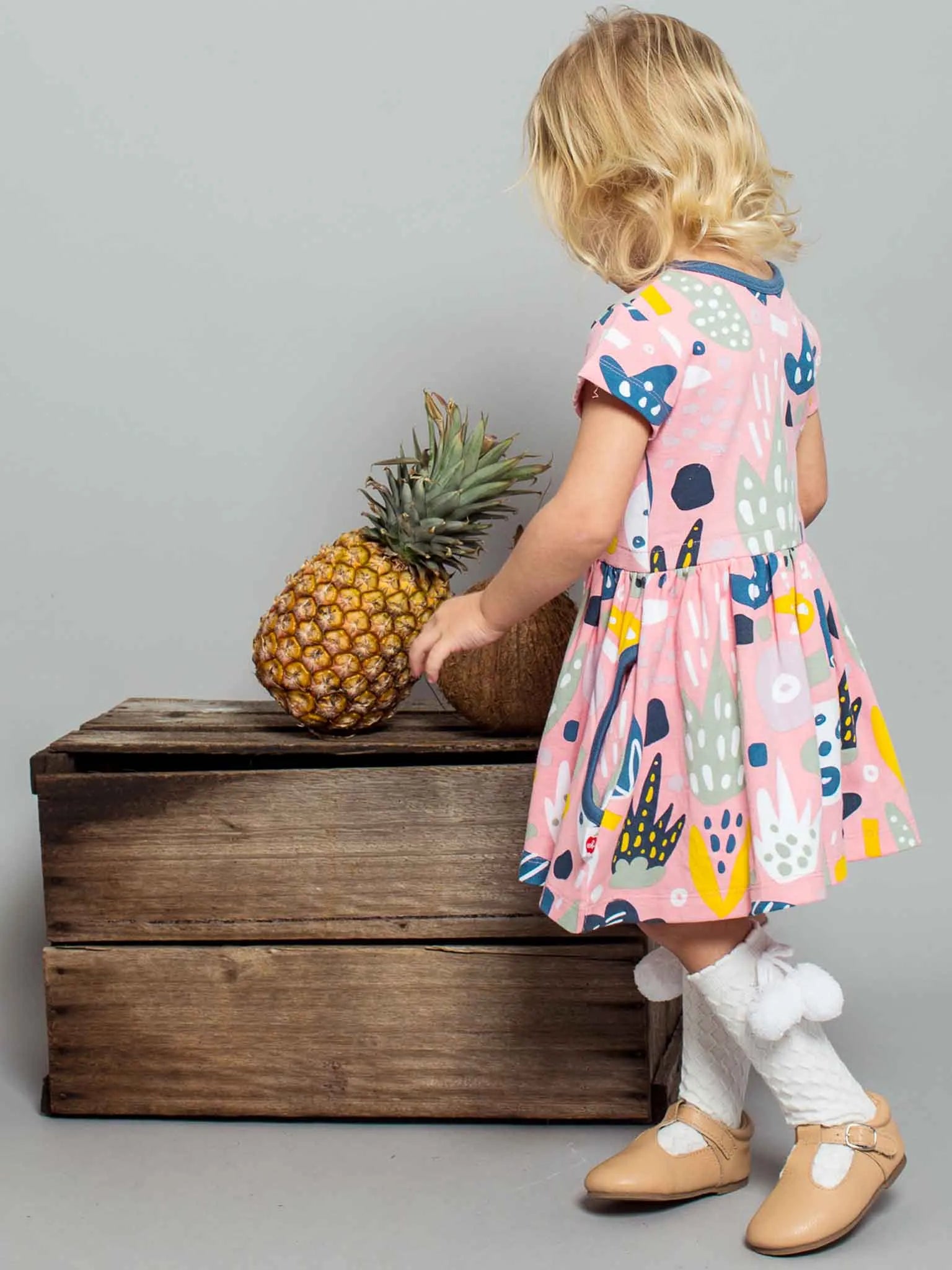 A little girl with blonde hair standing and wearing a pink jungle print dress and white pom pom socks