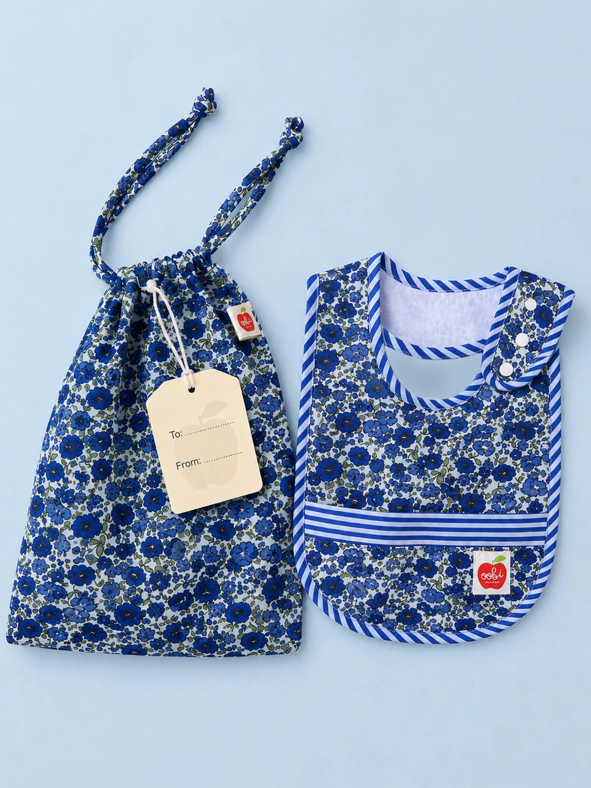 A matching baby gift set featuring a drawstring pouch and a bib in a blue and white floral print, displayed on a light blue background. The bib has a striped neck and waist trim and a small red apple logo patch.
