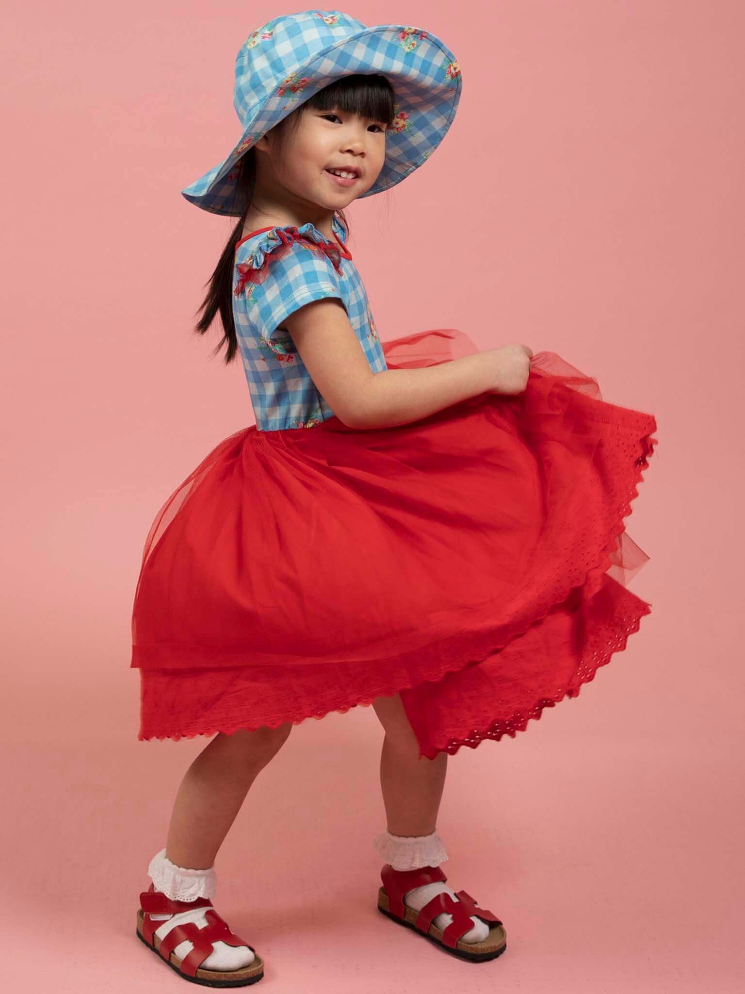 Young girl in a blue Floral Gingham Ponytail Hat and top, red tulle skirt, white socks, and red sandals stands against a pink background.